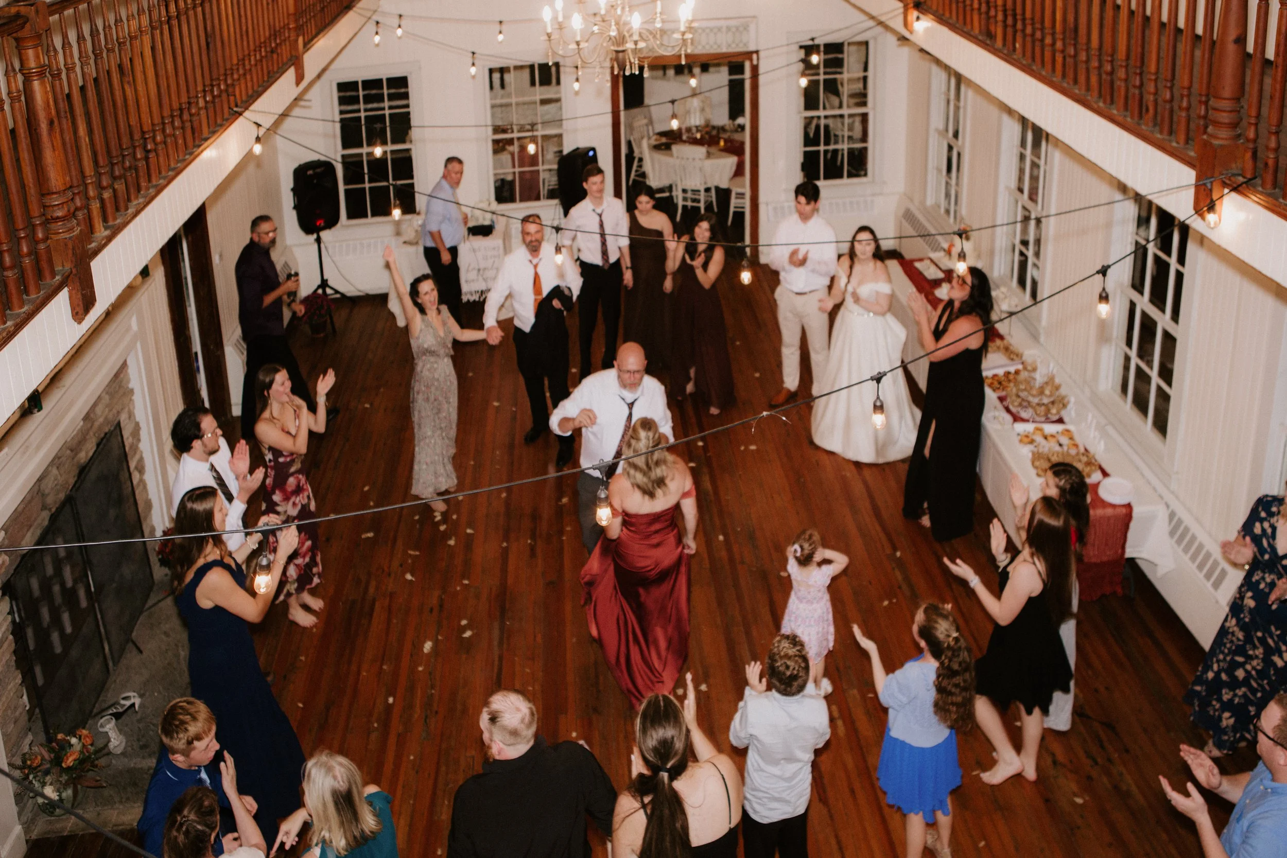 Guests dancing at a wedding reception in a spacious hall with wood floors, string lights, and large windows.