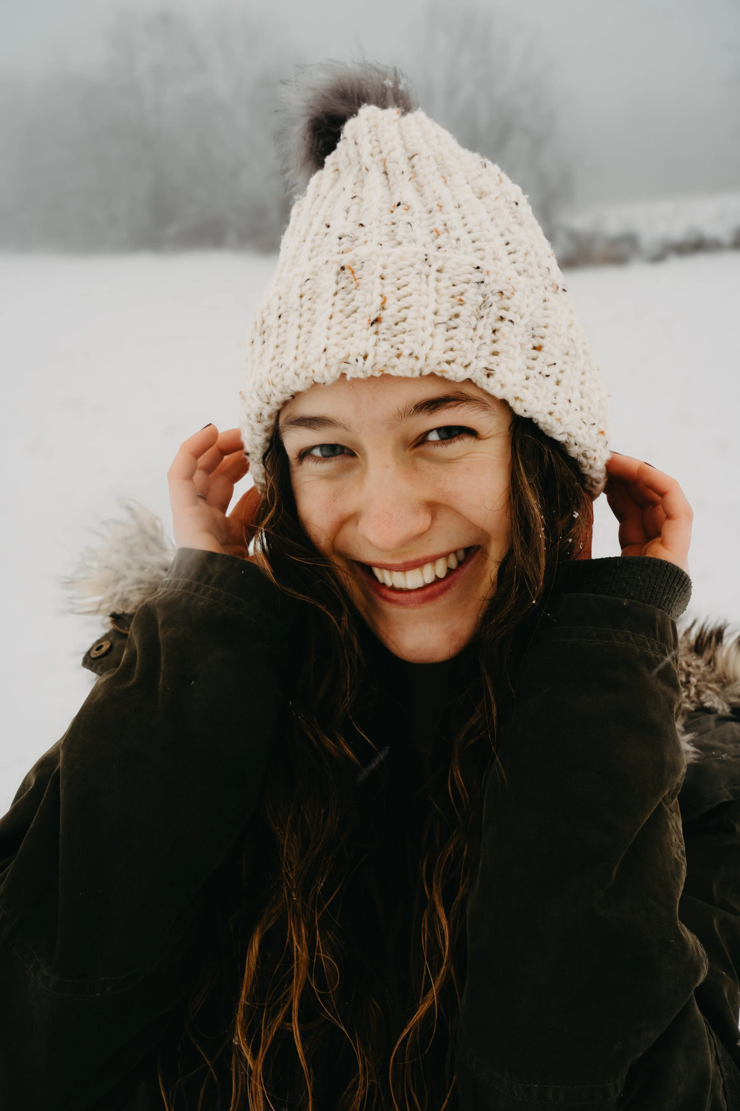 Close-up of smiling young woman wearing a knit hat and heavy winter coat in snowy outdoor setting.