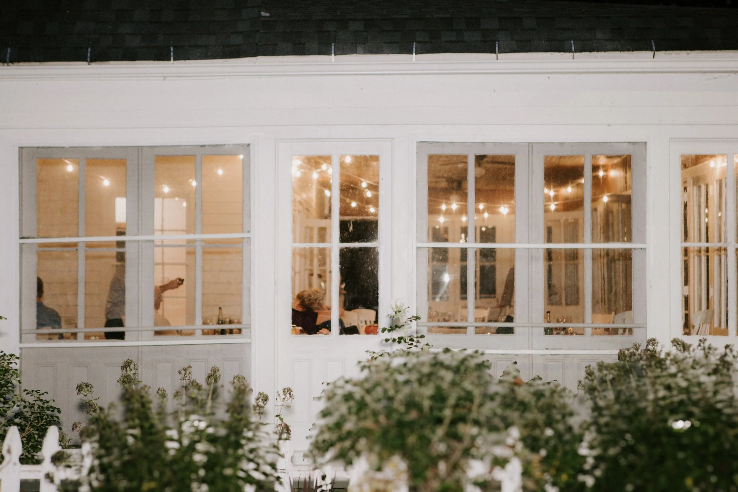 People gathered inside a house with large windows, sitting at tables and standing, illuminated by string lights hanging from the ceiling, viewed from outside through a garden.