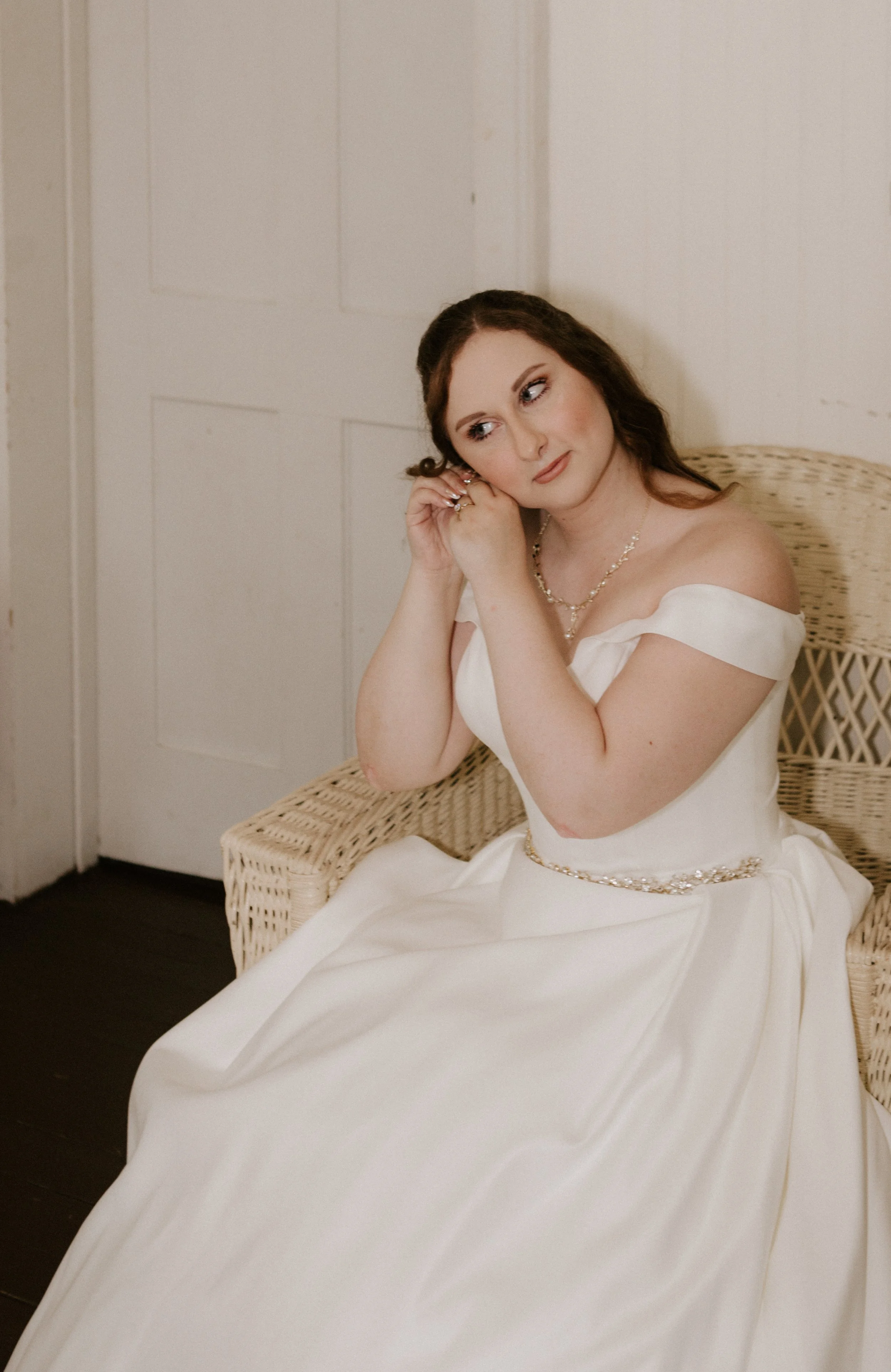 A bride sitting on a wicker chair, wearing an off-shoulder white wedding gown, earrings, and a necklace, adjusting her earring with a contemplative expression.