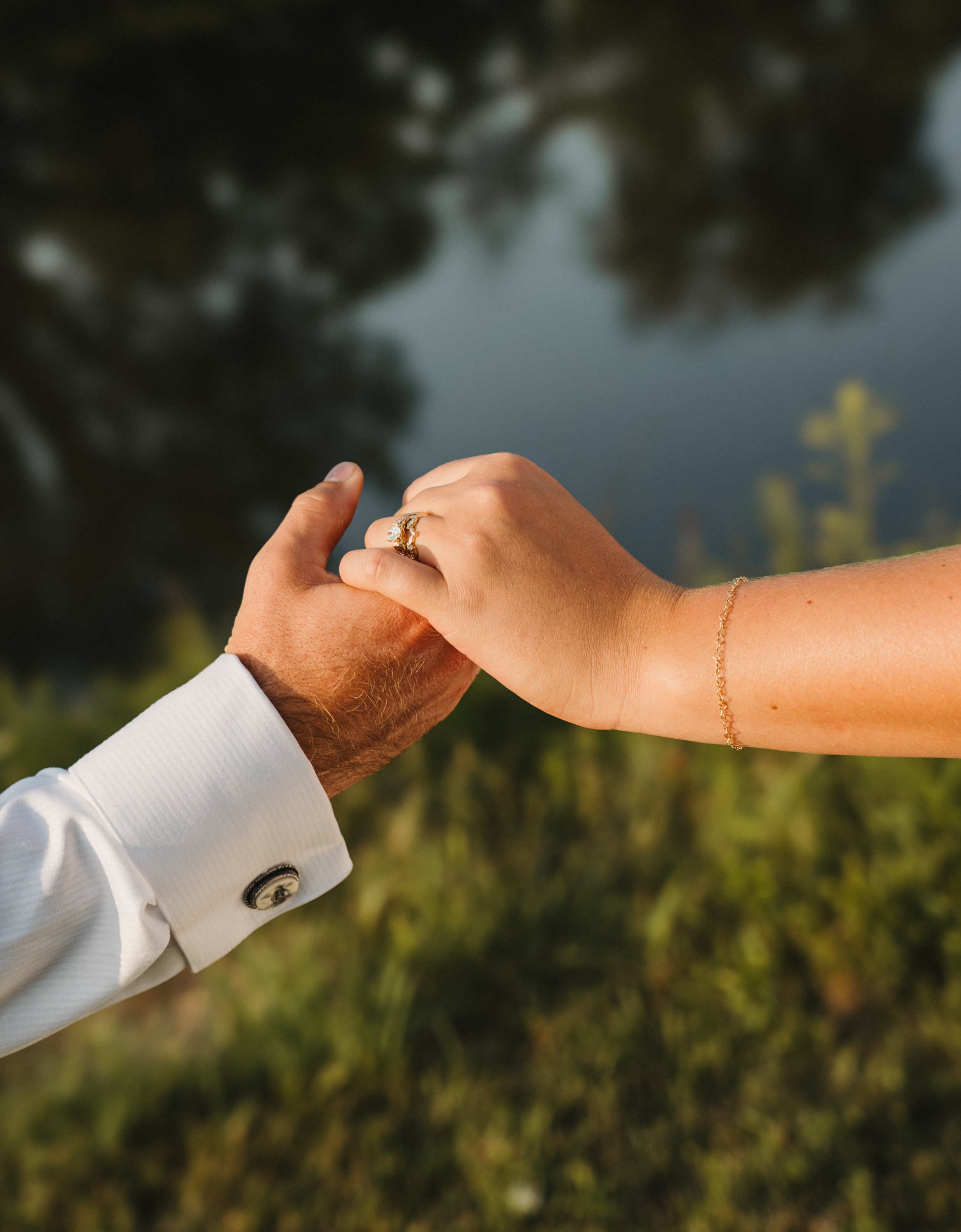 Close-up of two hands holding each other near a body of water with trees in the background. One hand appears more masculine, and the other more feminine, both adorned with jewelry.