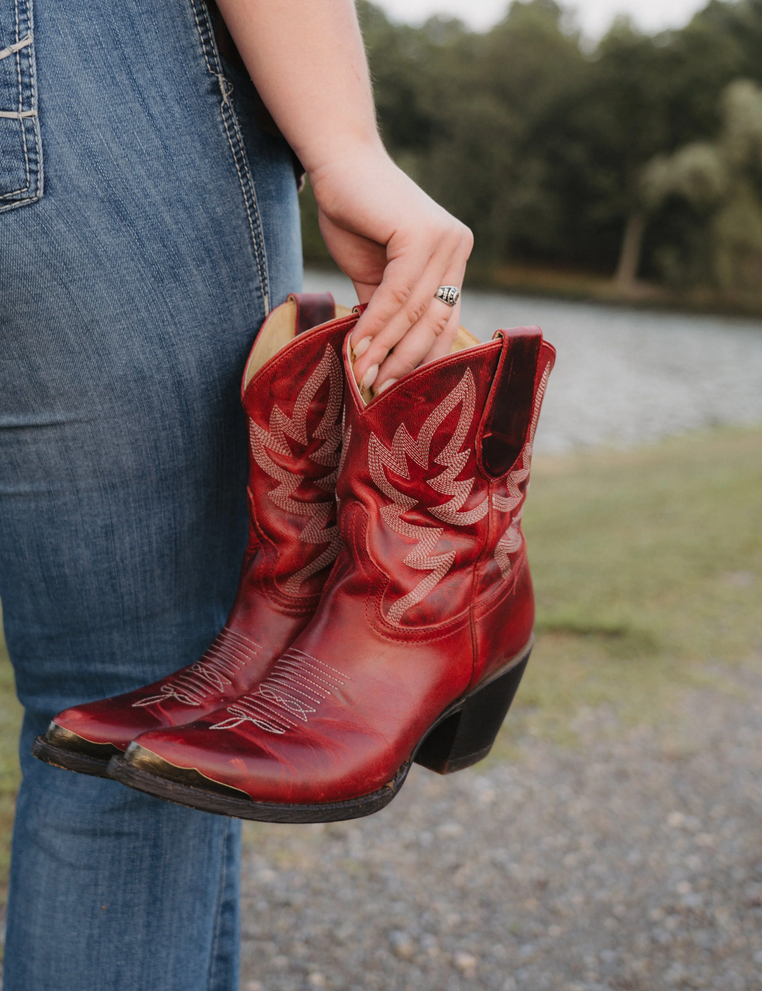 Person holding a pair of red cowboy boots with white embroidery, standing outdoors near a body of water with trees in the background.