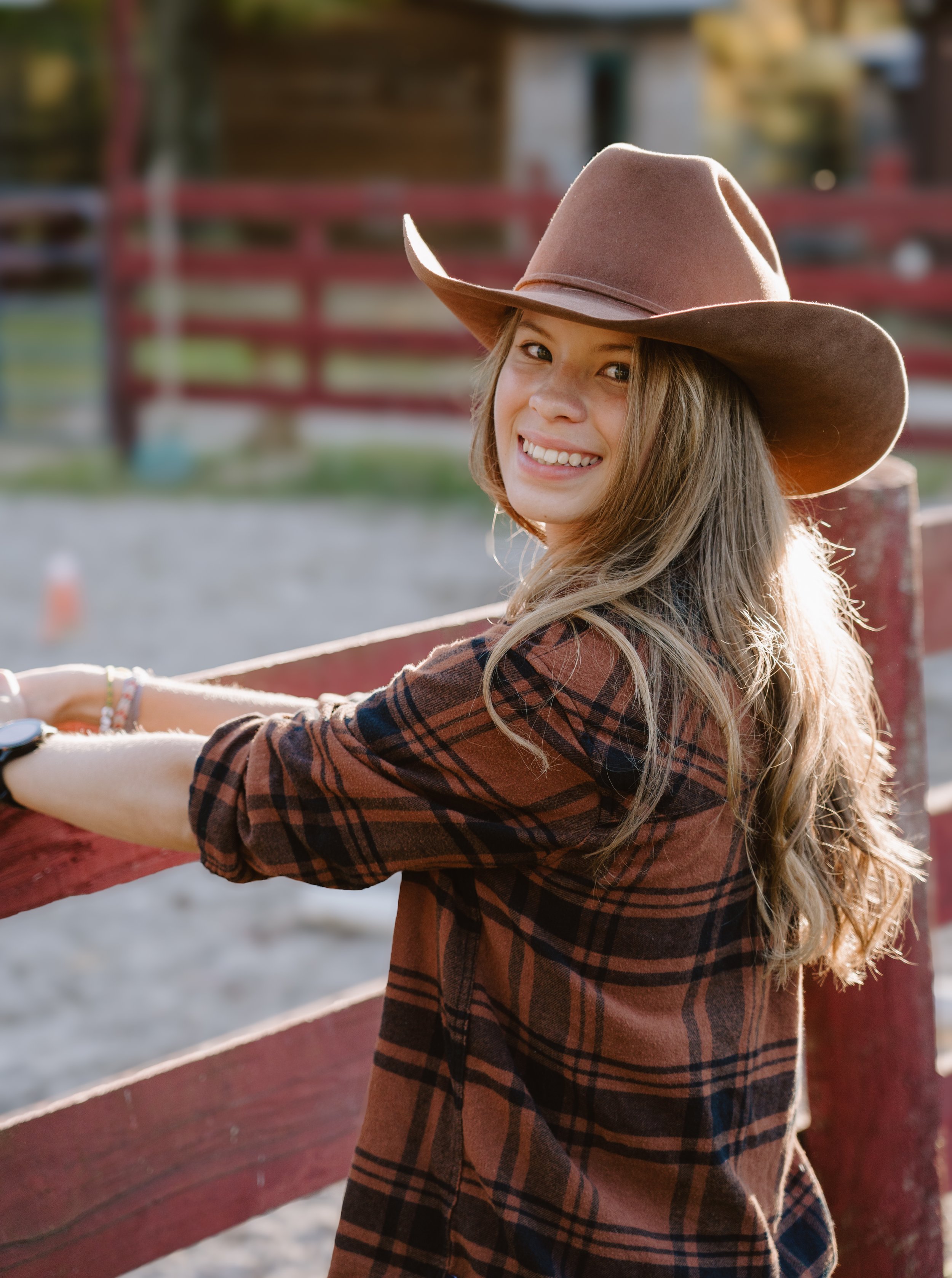 A young woman wearing a brown cowboy hat and a plaid shirt, smiling and leaning on a red wooden fence outdoors during daylight.