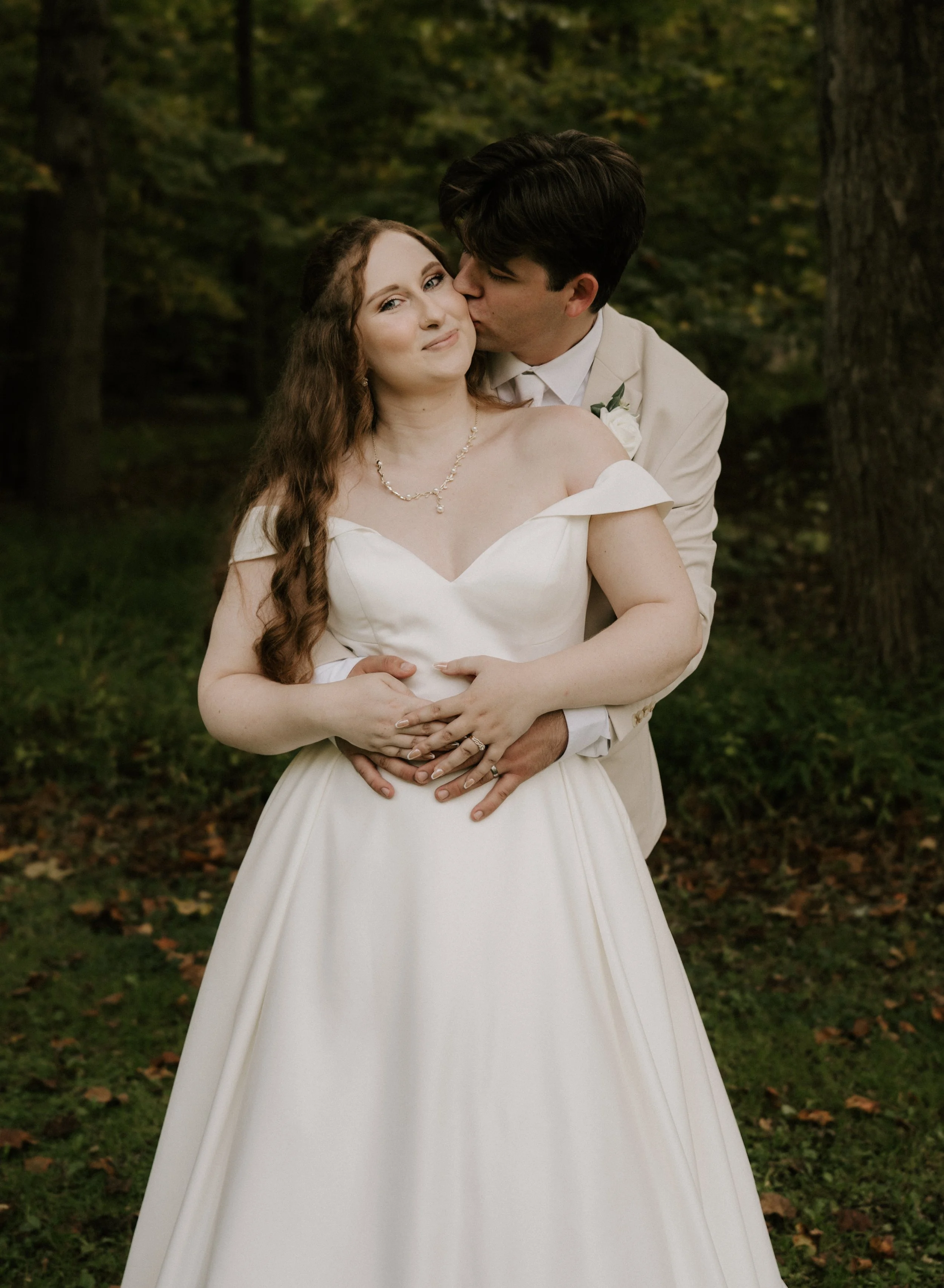 A newlywed couple in wedding attire posing outdoors in a forest setting, with the groom kissing the bride's cheek and holding her waist, while the bride looks at the camera.
