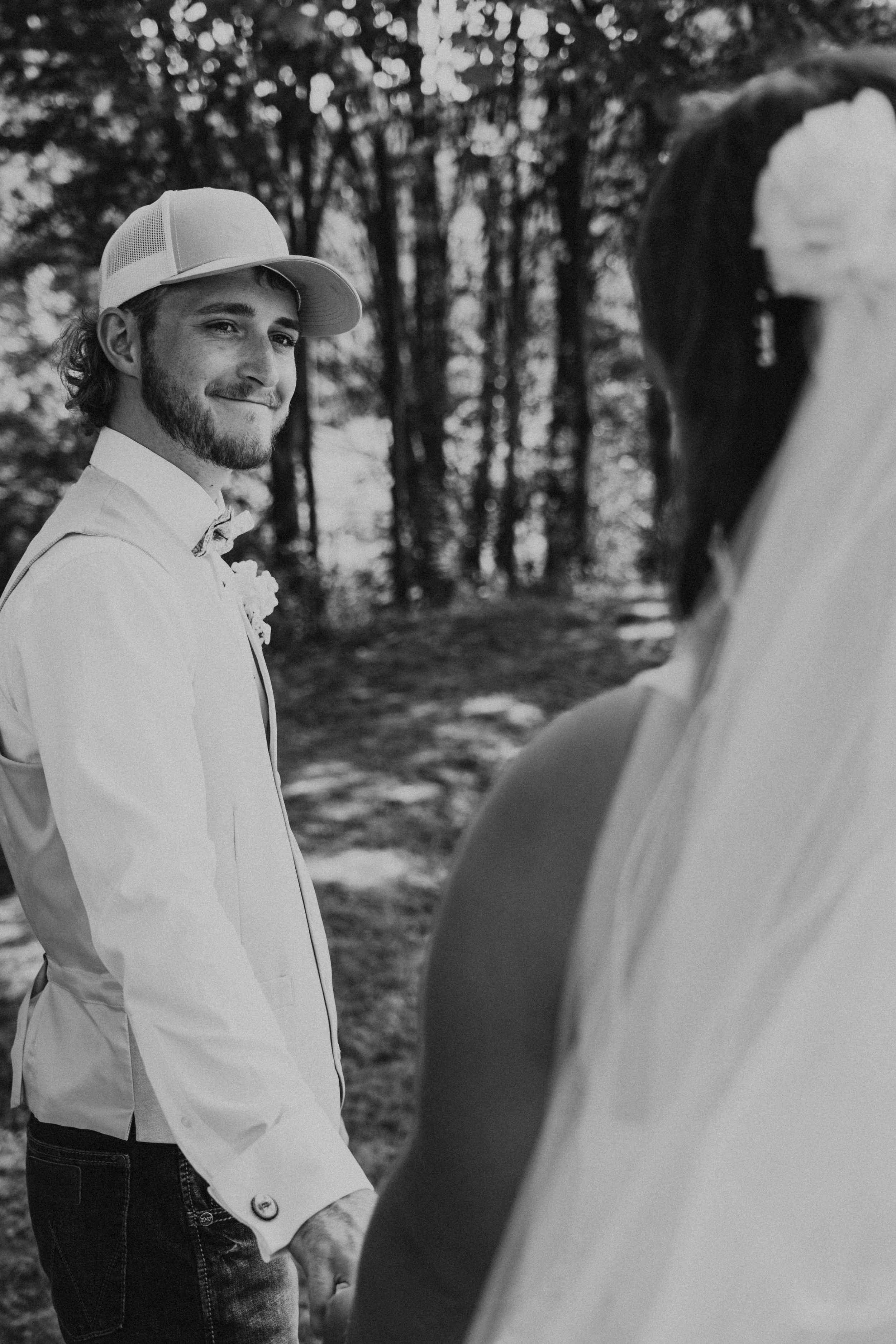 A man in a white shirt, vest, and baseball cap smiling and holding hands with a woman in a wedding dress outdoors in a forest setting.