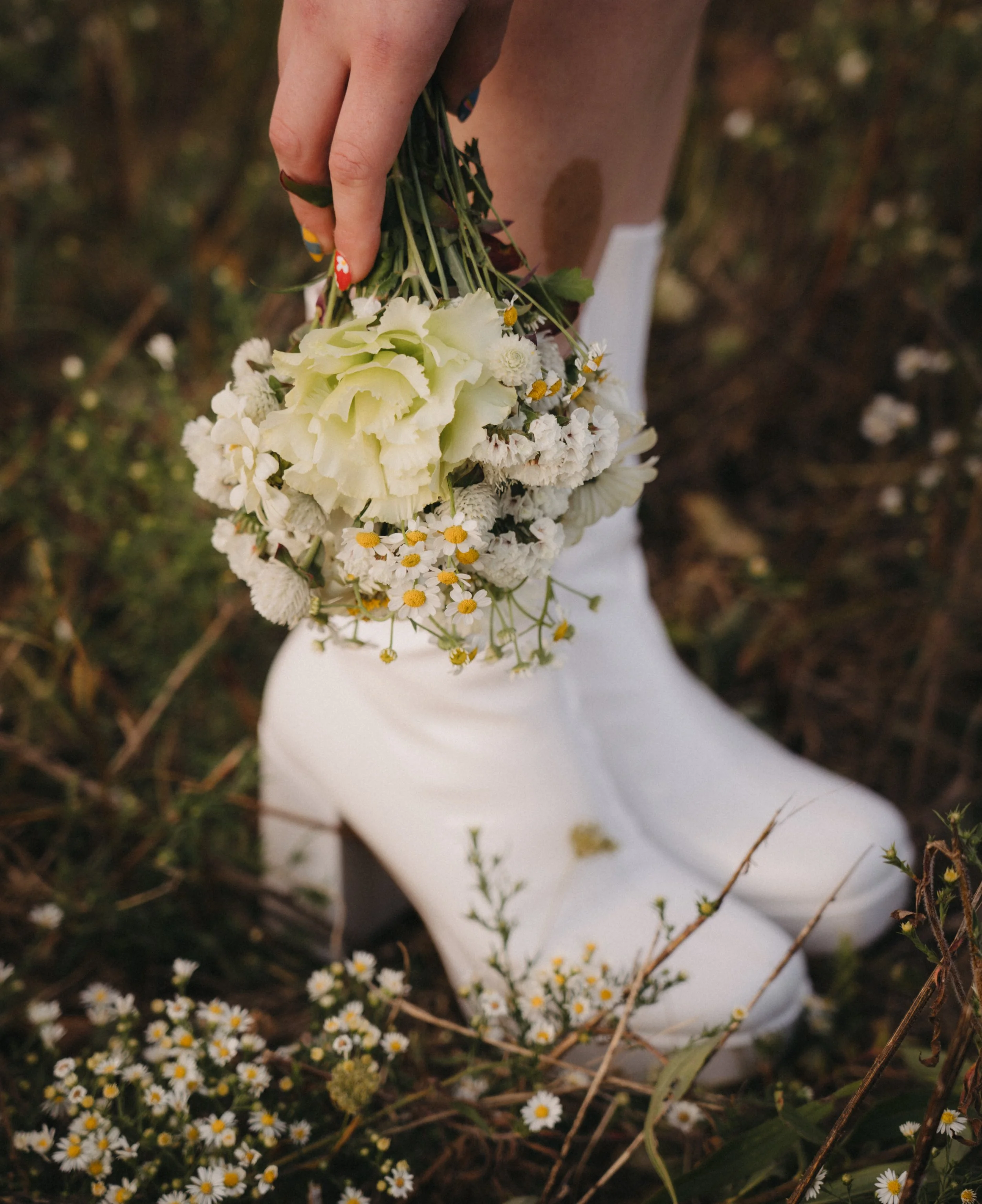 Person wearing white cowboy boots holding a bouquet of white flowers in an outdoor setting with wildflowers and grass.