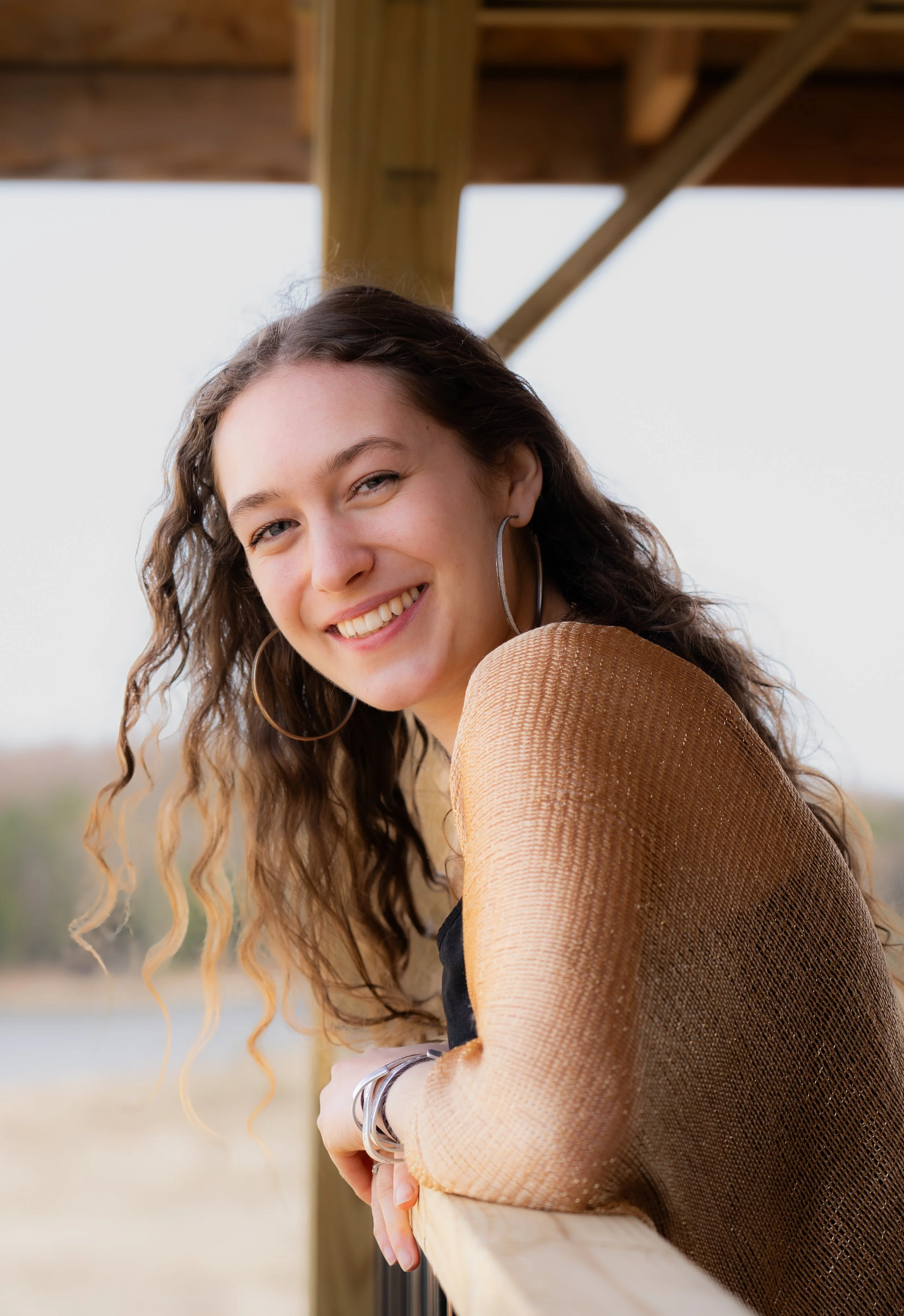 Young woman with curly brown hair leaning on a wooden railing outdoors, smiling at the camera, wearing large hoop earrings and a tan textured jacket.