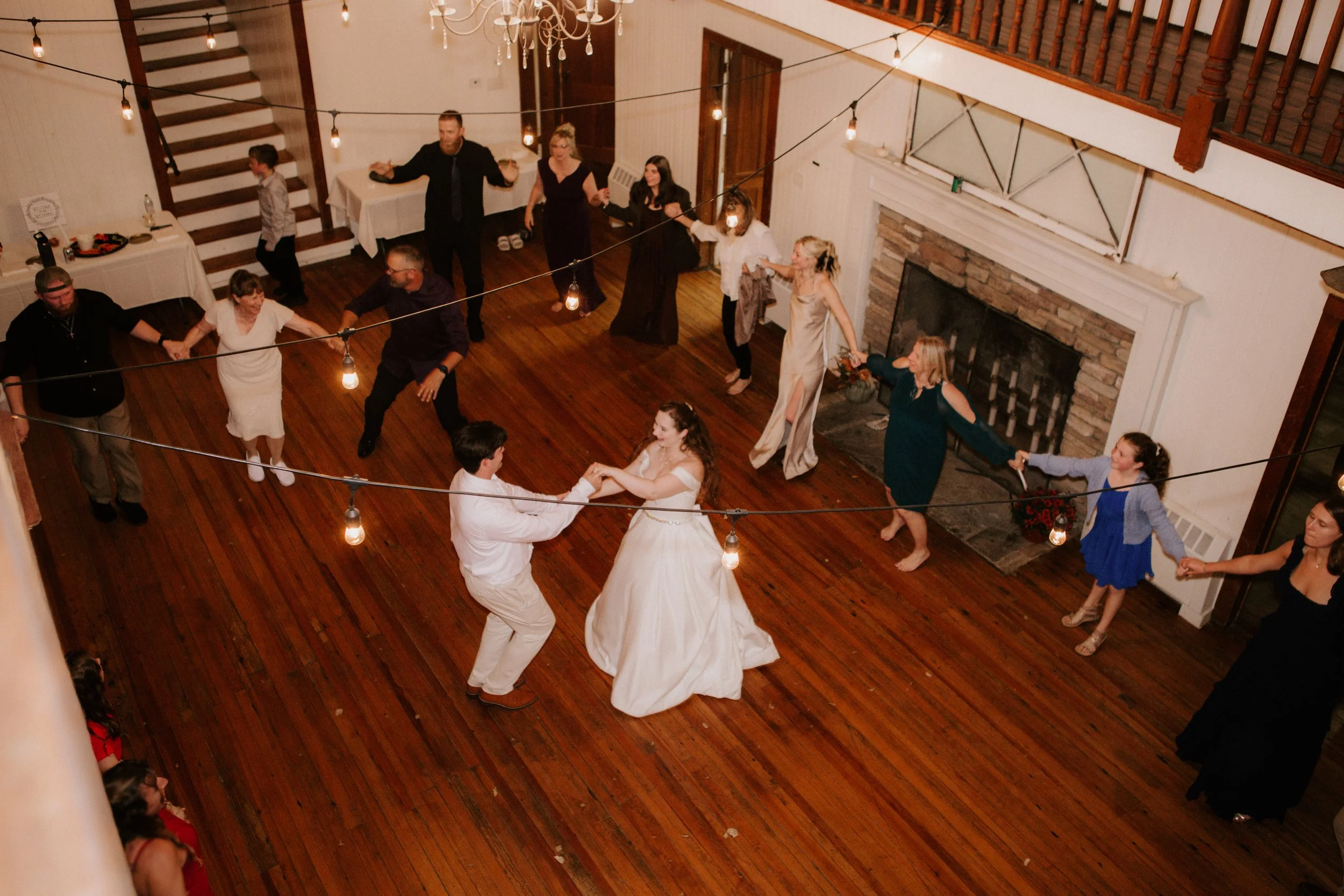 A bride and groom dancing and holding hands in the center of a wooden-floored room with string lights overhead at a wedding reception, surrounded by guests holding hands and dancing in a circle.