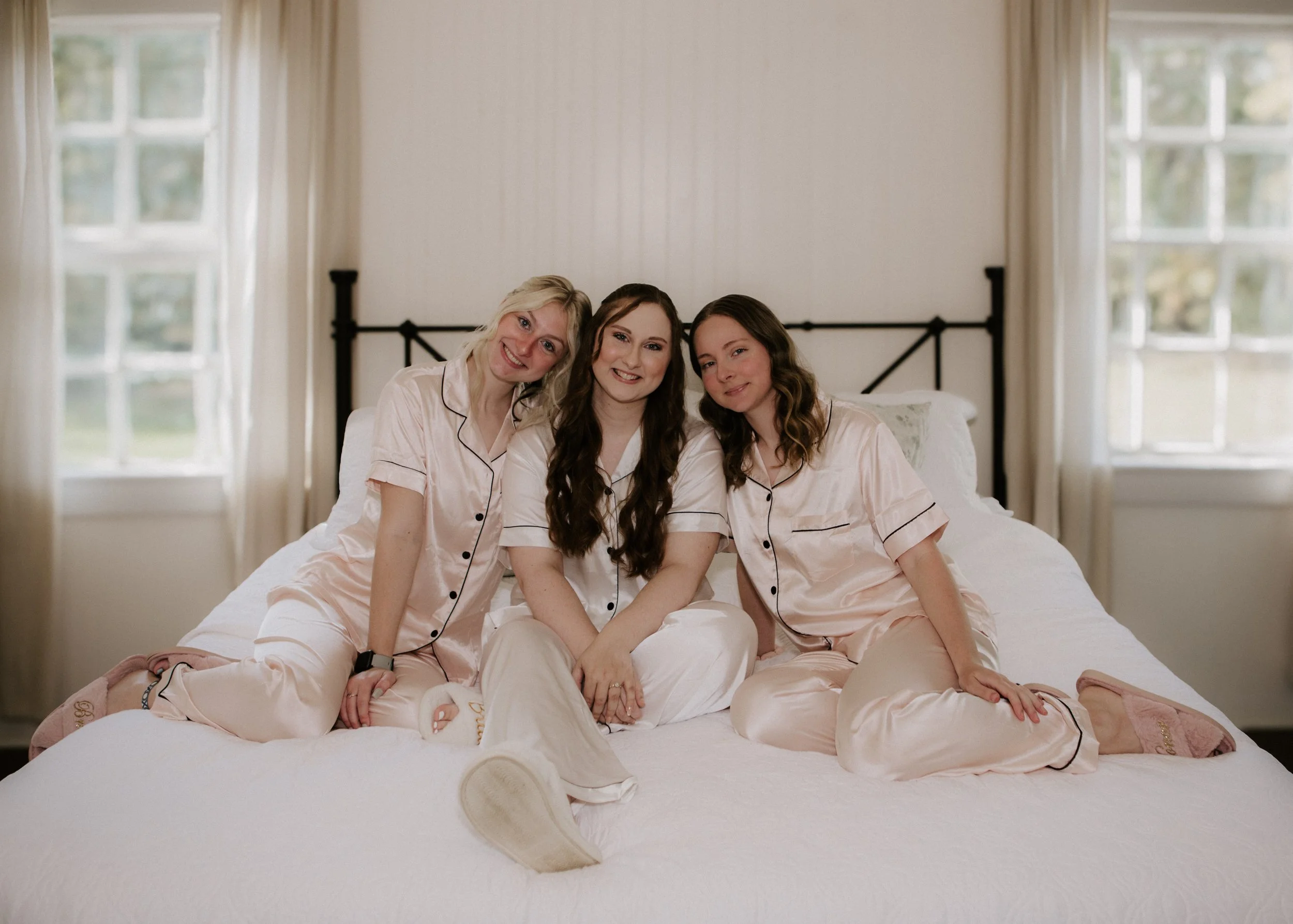 Three women sitting on a bed in pajamas, smiling at the camera in a well-lit bedroom with large windows and cream curtains.