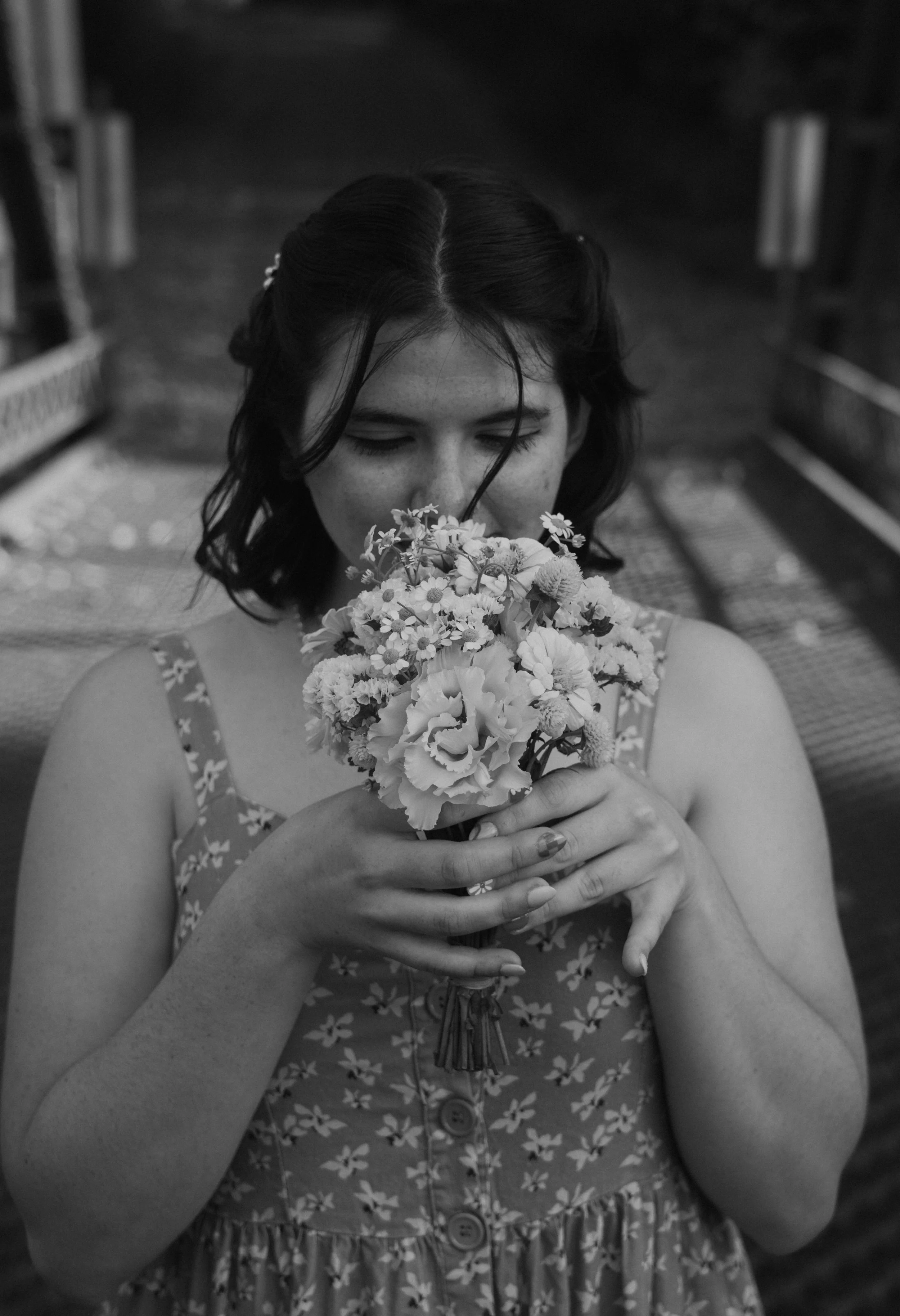 A young woman with dark hair and freckles holding a bouquet of flowers with both hands, her eyes closed and nose touching the flowers.