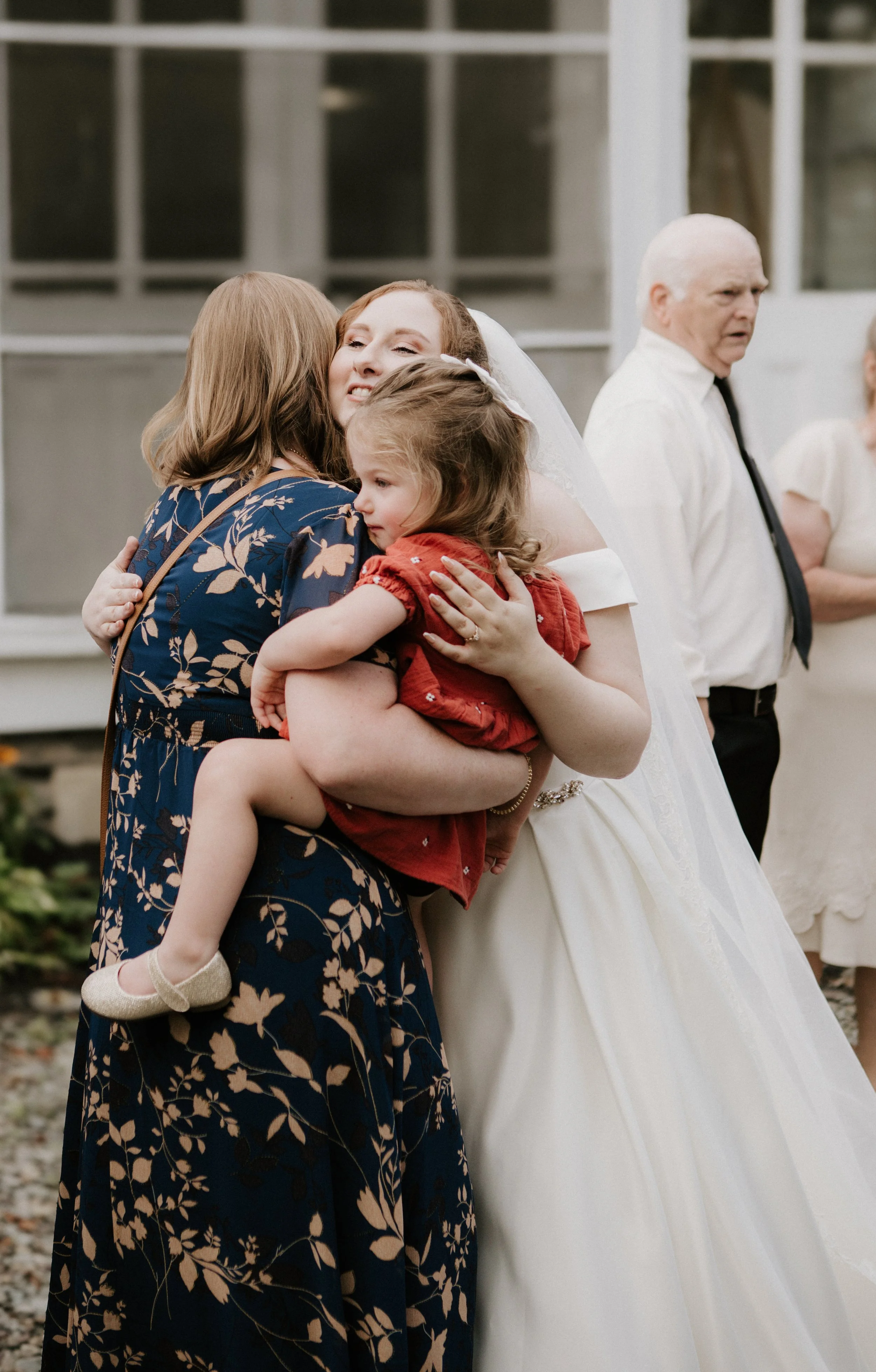 A bride hugging a woman and a young girl, with a man in a white shirt and black tie in the background at a wedding.