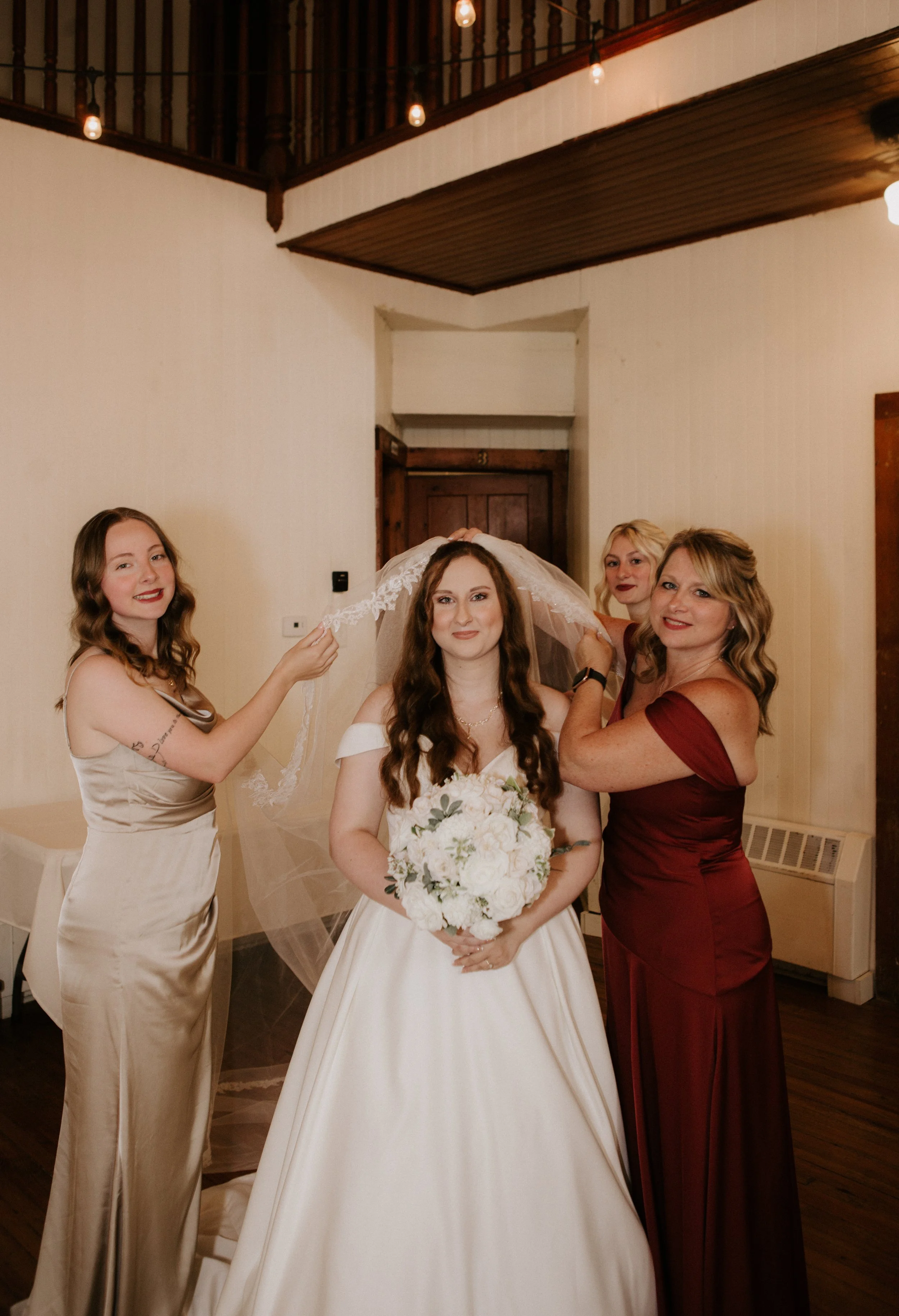 A bride holding a bouquet of white flowers with three women helping with her veil in a warmly lit room.