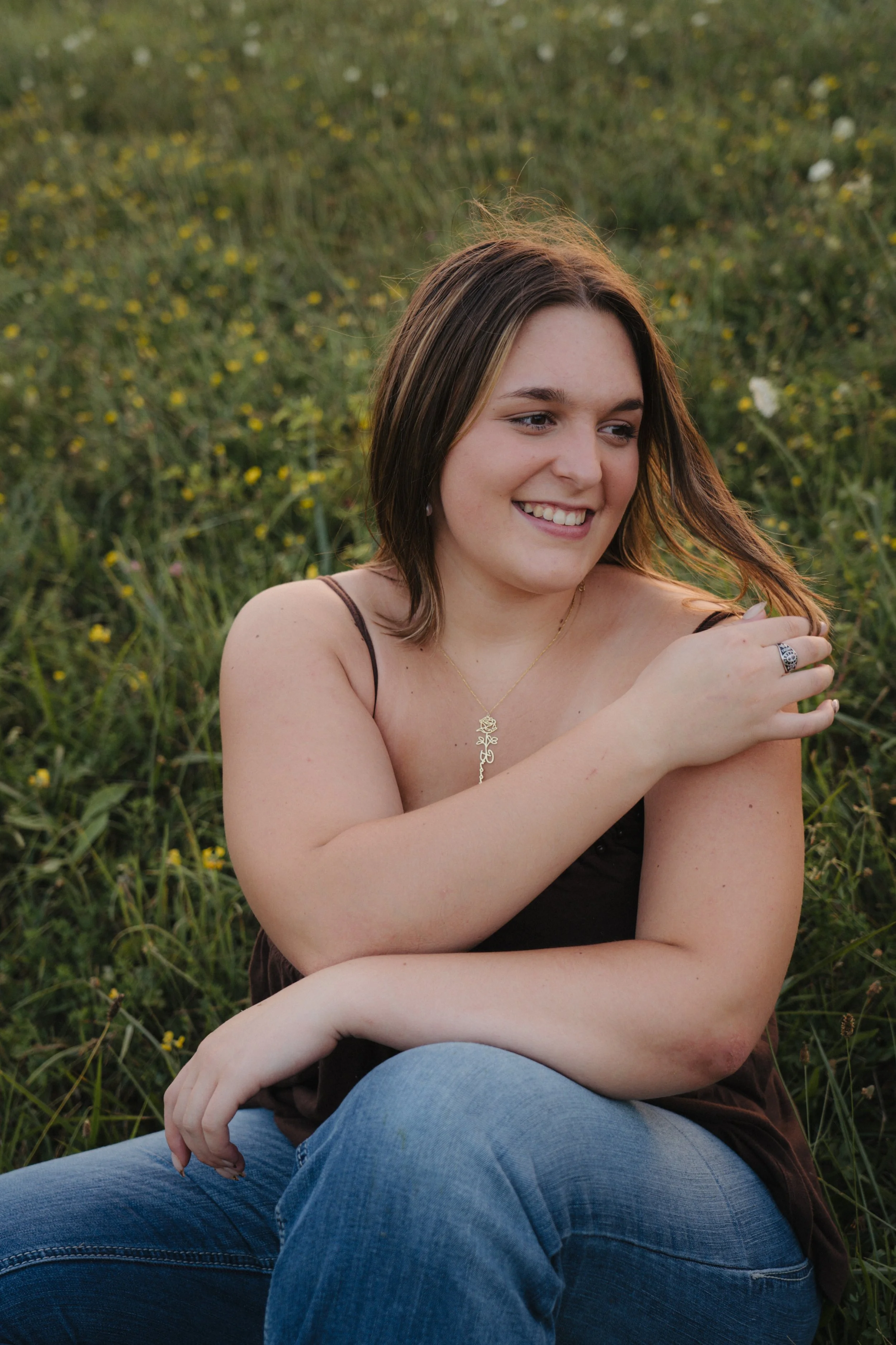 A young woman sitting in a grassy field with small yellow flowers, smiling and looking to the side, wearing a black tank top and blue jeans.