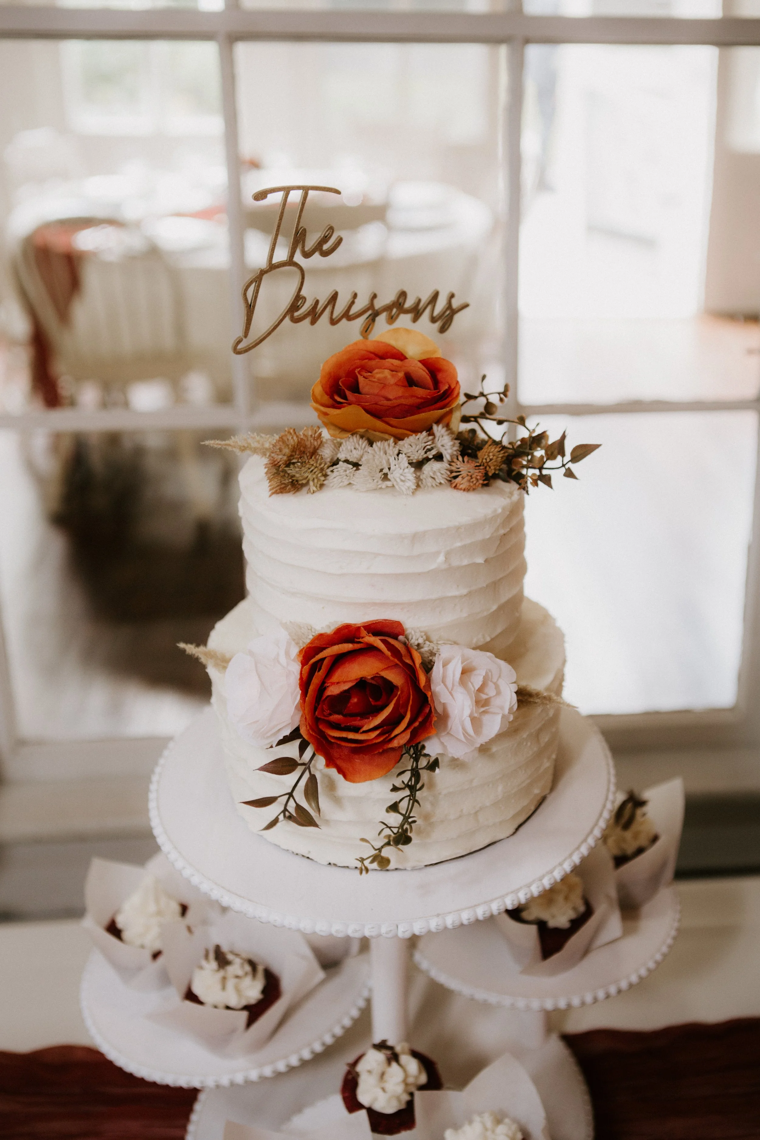 A two-tiered white wedding cake decorated with orange and white roses, greenery, and a gold custom cake topper reading 'The Densons'. The cake is on a white stand with cupcakes around it, placed near a glass door with a blurred sofa and window in the