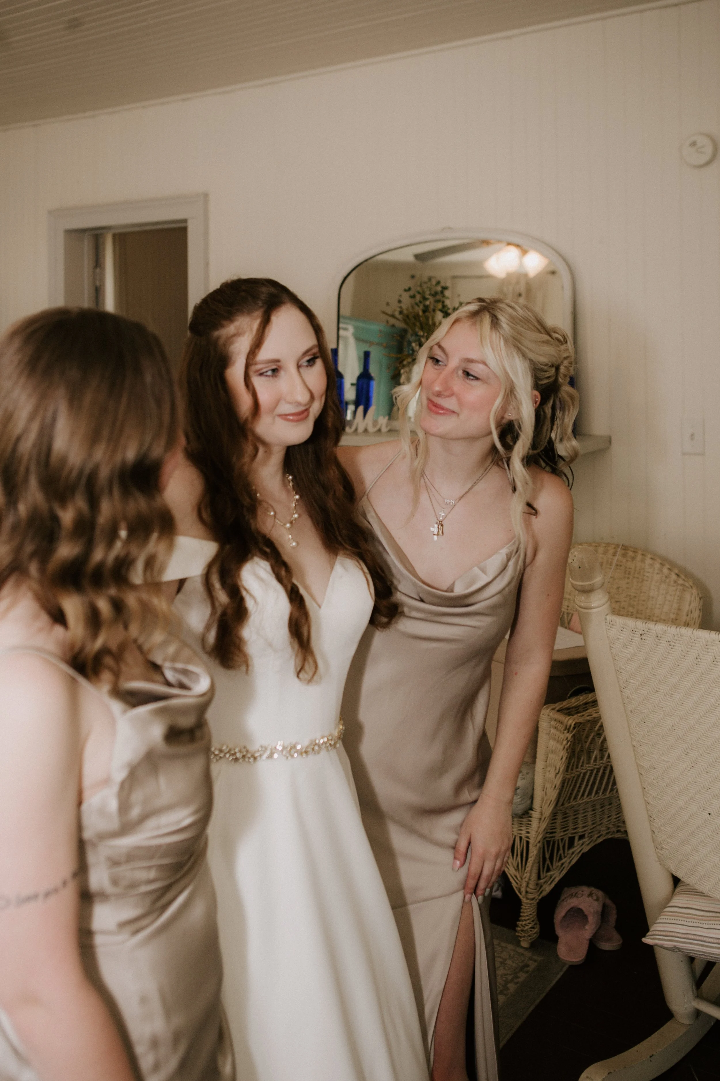 A bride with long brown hair in a white wedding dress standing with two bridesmaids in beige satin dresses in a room with white walls and wicker furniture.