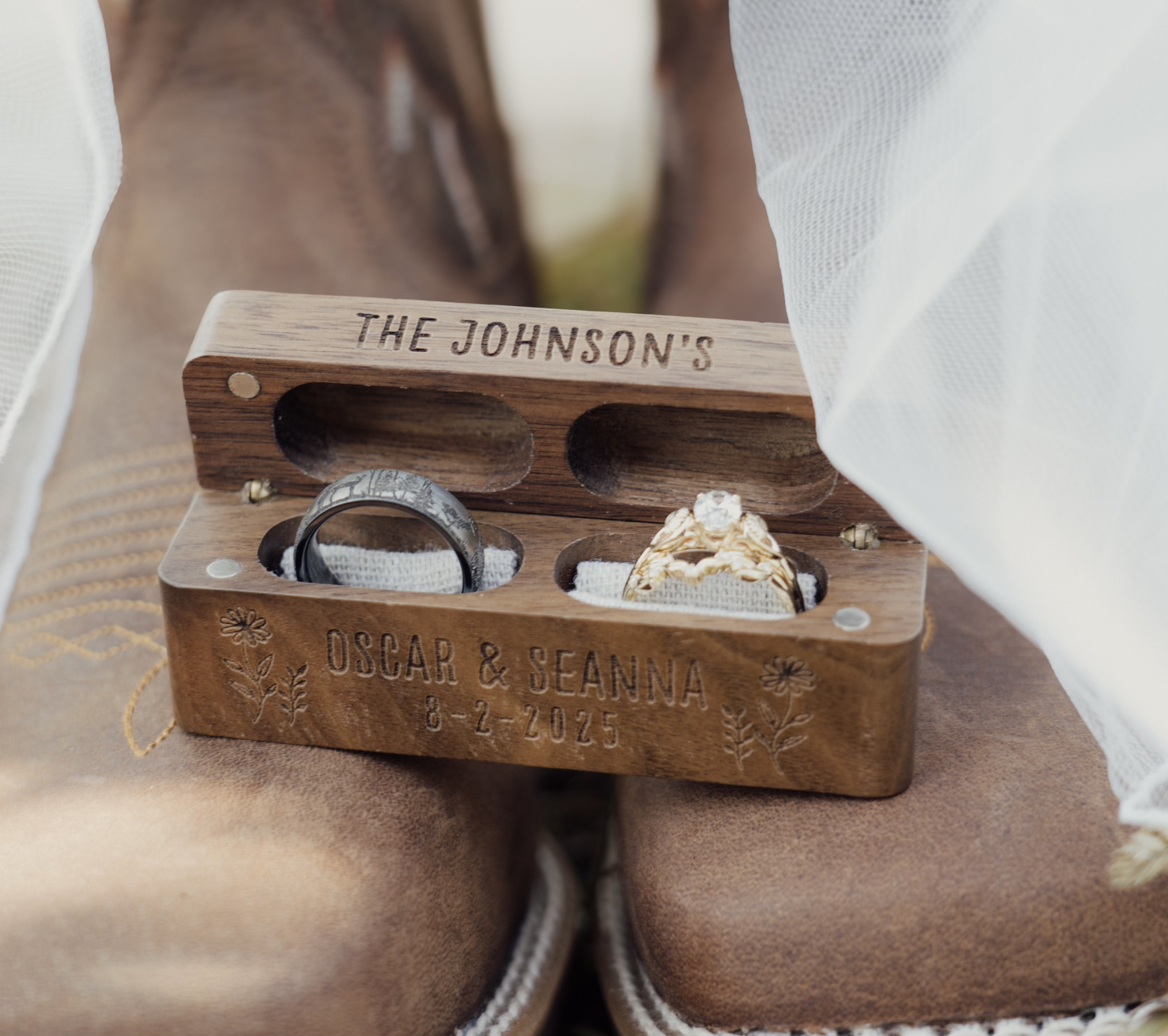 Wooden box engraved with the names Oscar and Beanna, the date 8-2-2025, and floral decorations. Inside the box are two wedding rings, one with a diamond, resting on fabric.