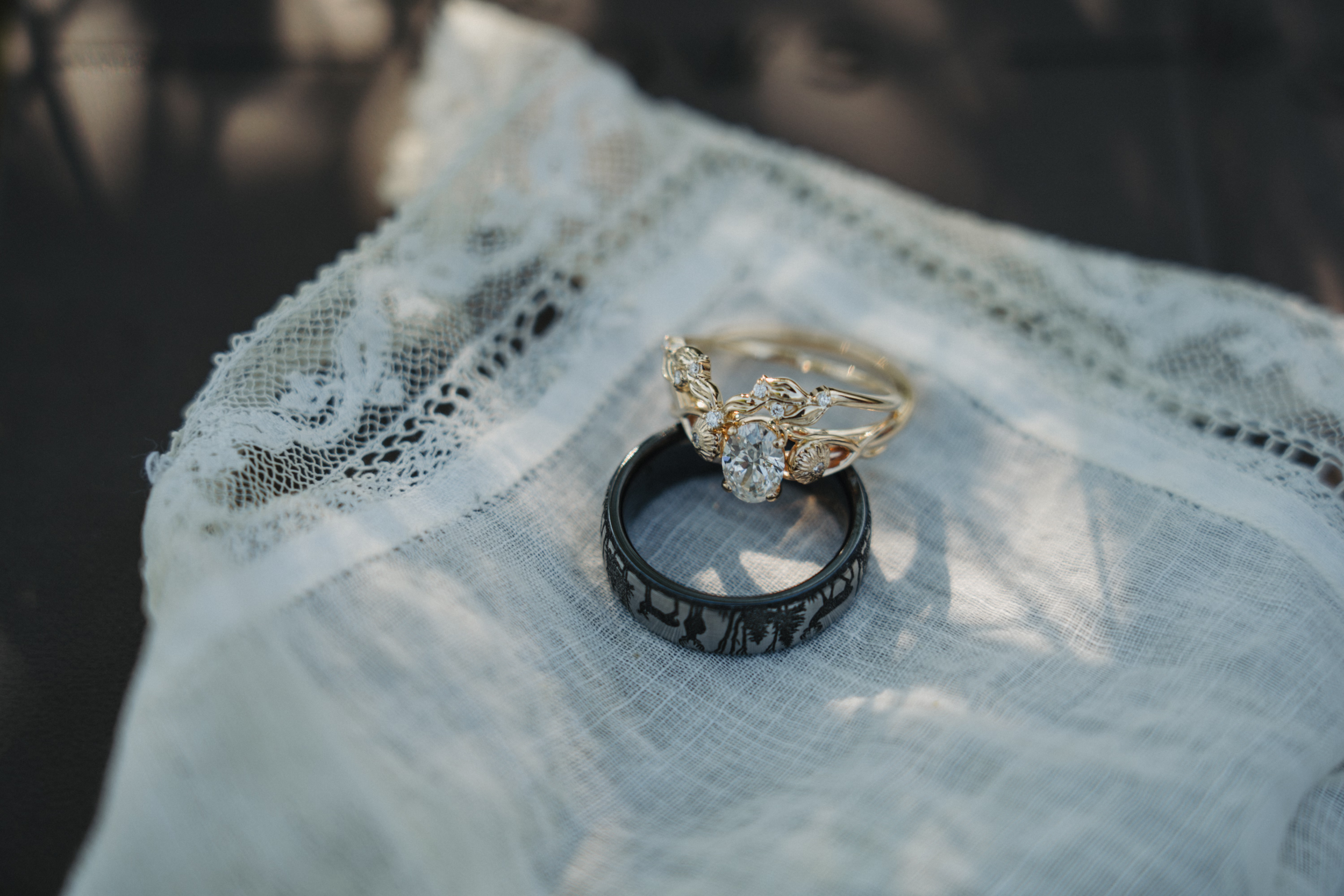Close-up of two wedding rings, one with a large central diamond and intricate gold detailing, and the other a plain black band with a pattern. The rings are placed on a piece of white lace fabric.