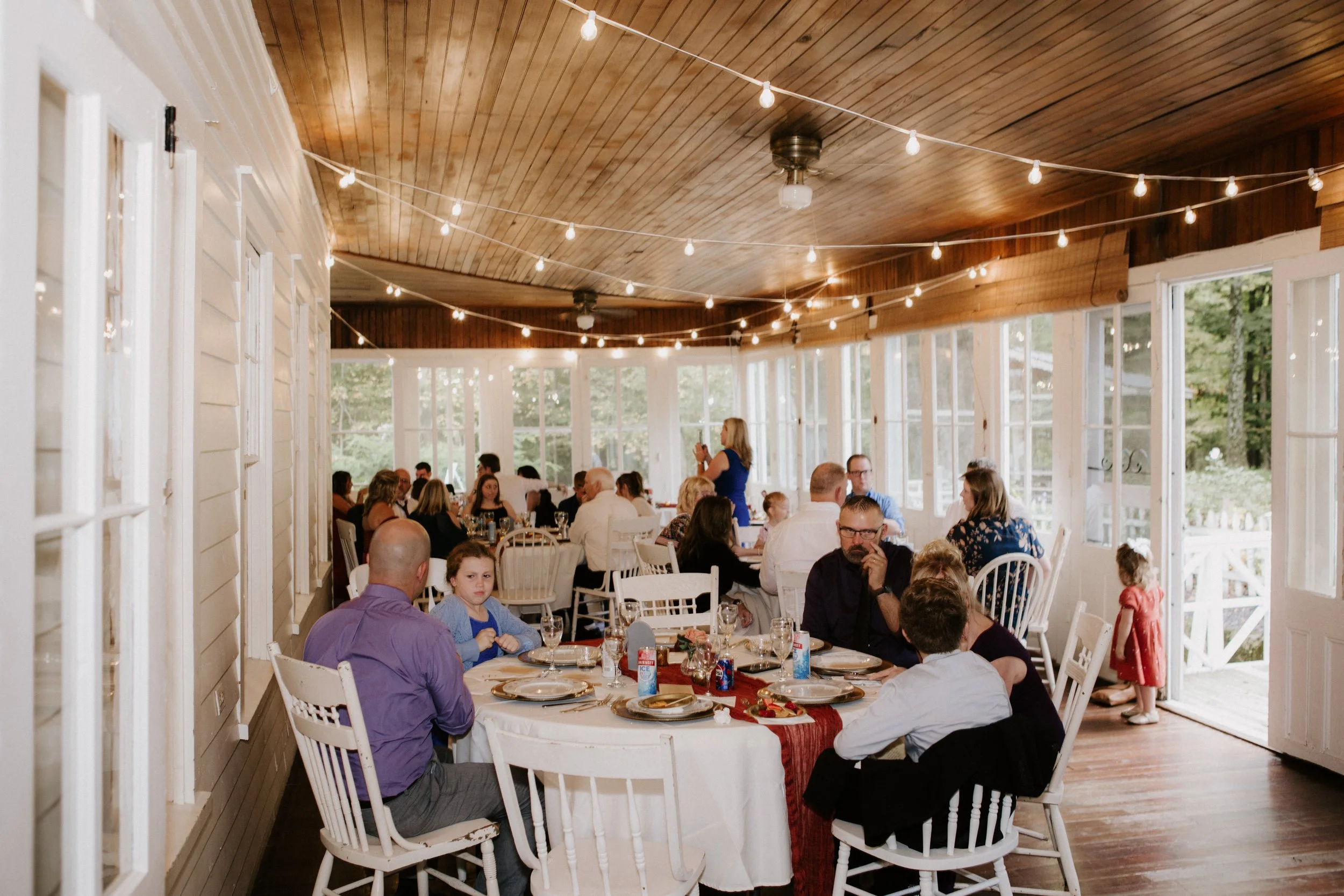 Indoor family gathering with multiple round tables set with plates, glasses, and decorations. String lights hang from the wooden ceiling. People are seated, talking, and a woman stands and speaks. Child in a red dress stands near open door with a whi