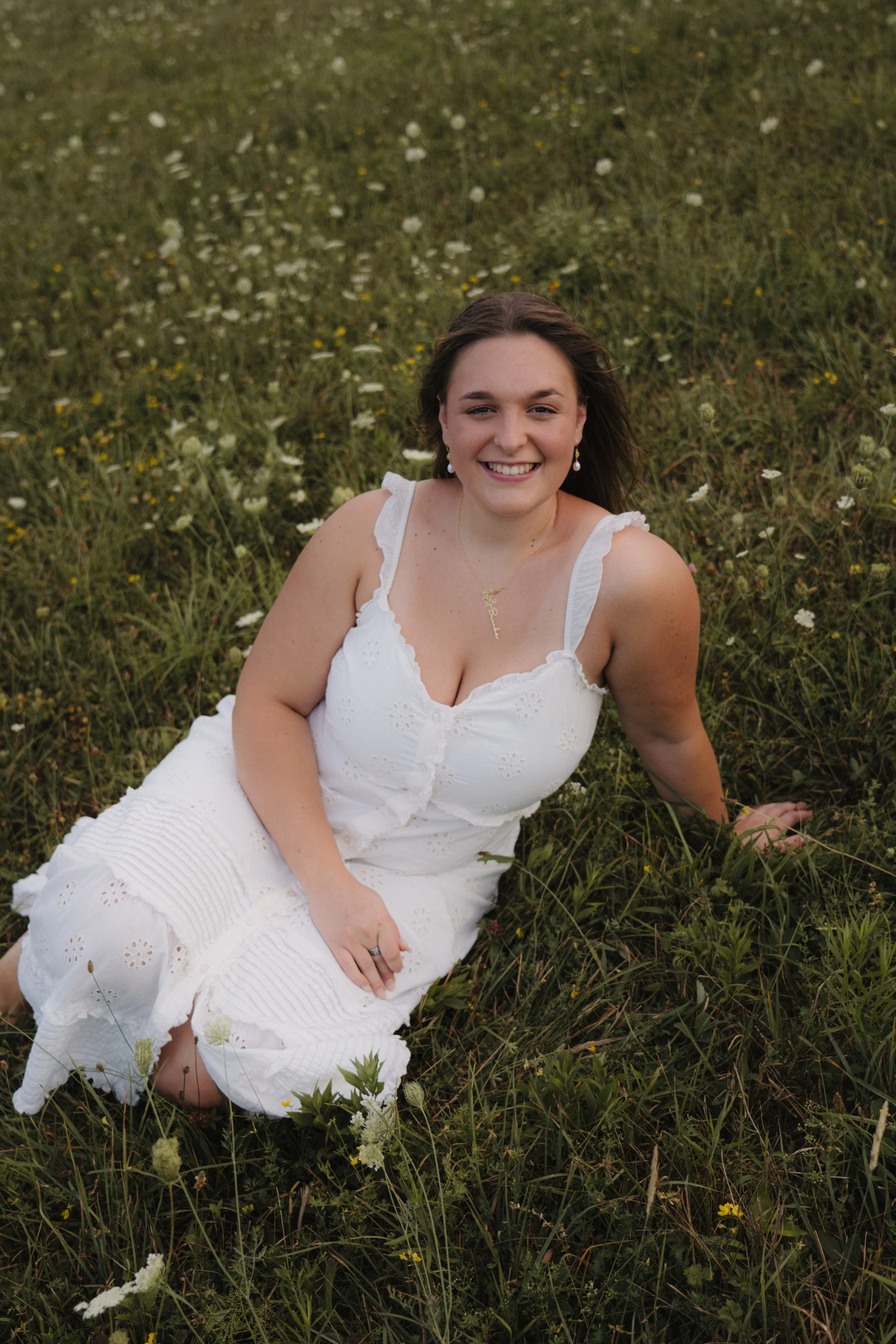 A woman in a white dress with eyelet details, sitting on grassy land in a field of wildflowers, smiling at the camera.