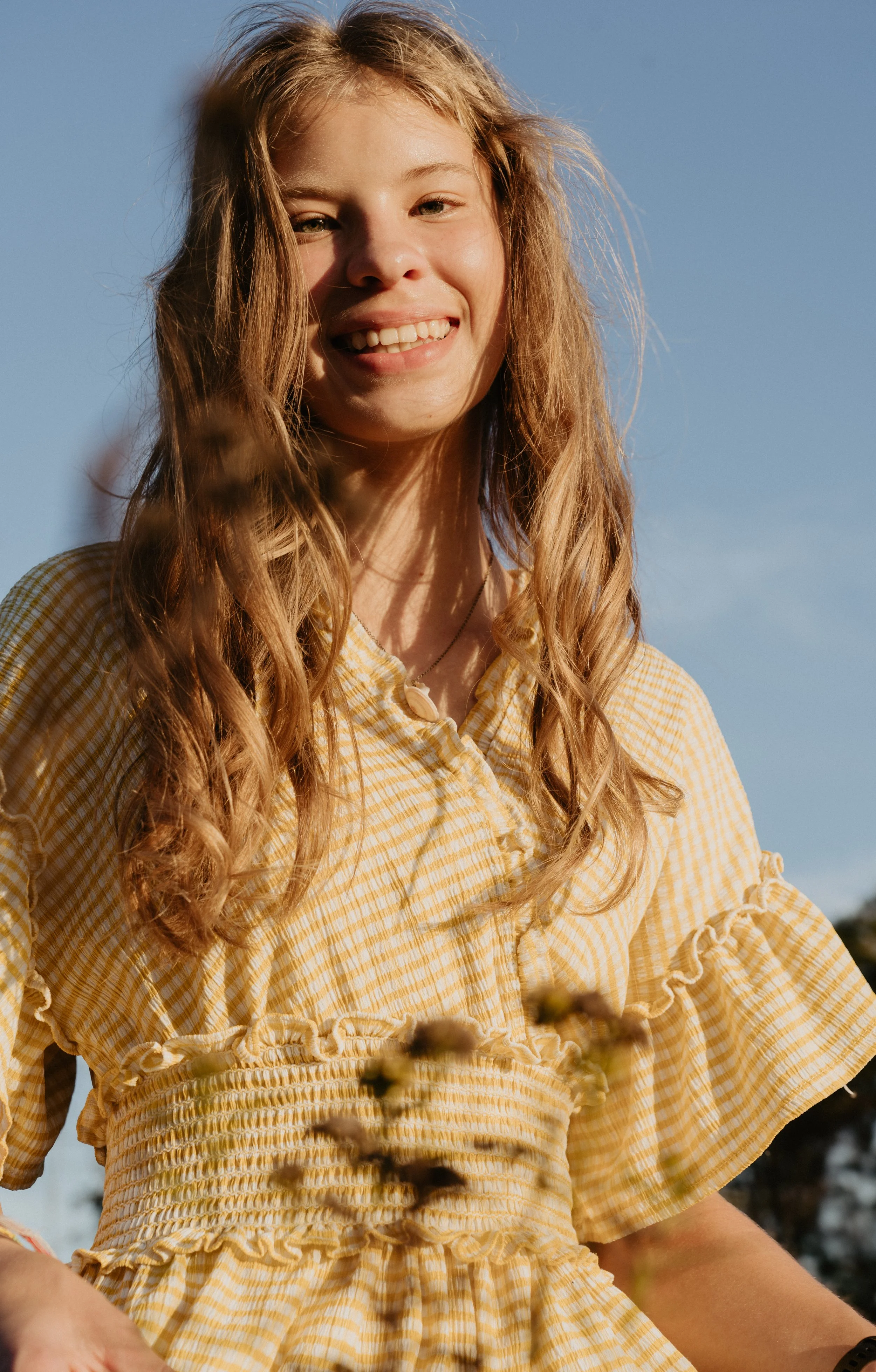 A young woman with long, wavy blonde hair smiling outdoors against a blue sky, wearing a yellow gingham dress with ruffled sleeves.