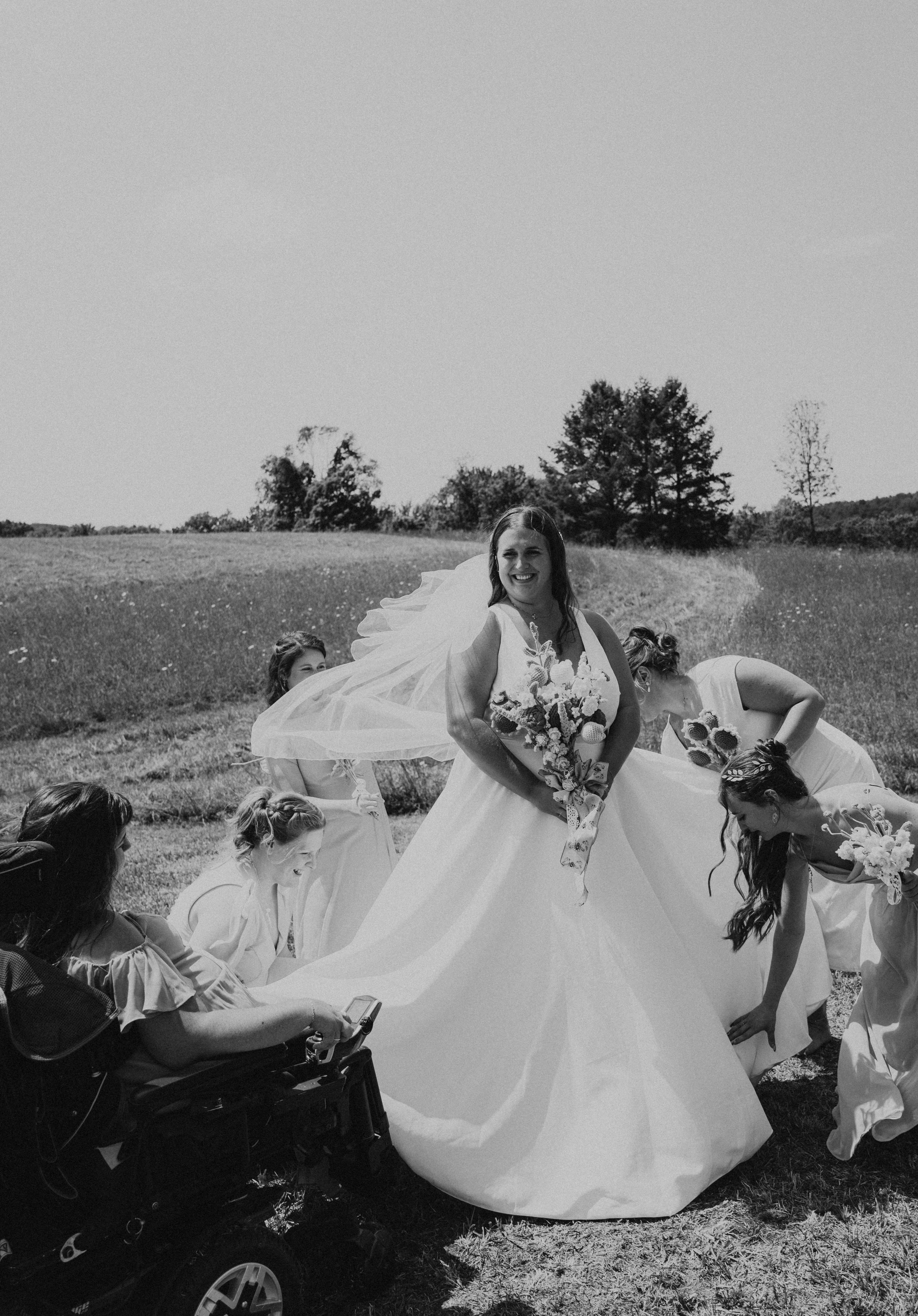 A black-and-white photo of a bride in a wedding dress holding a bouquet, surrounded by bridesmaids and a woman in a wheelchair, in an outdoor field with trees in the background.