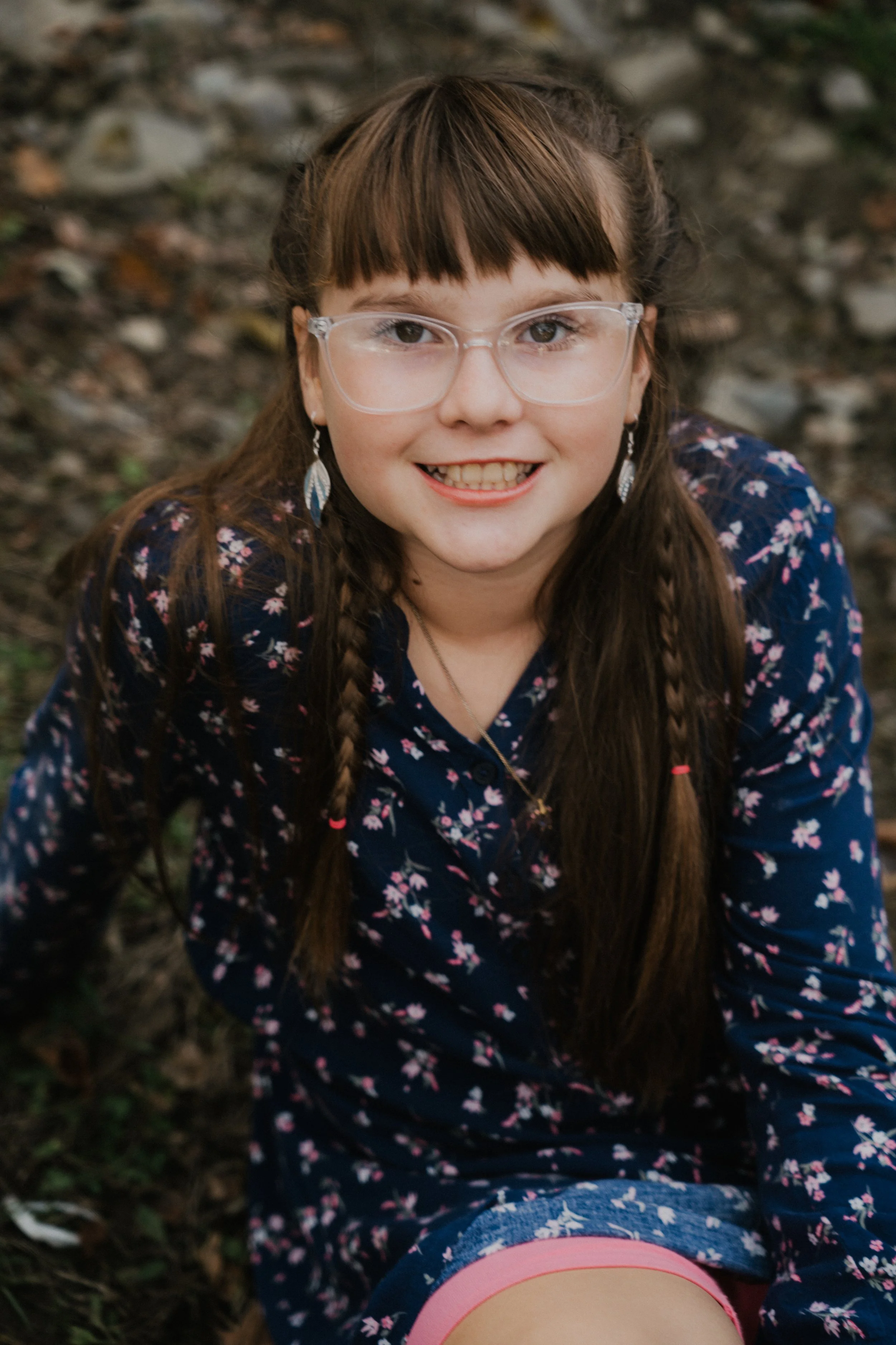A young girl with glasses, long brown hair in braids, and a floral navy dress, smiling outdoors.