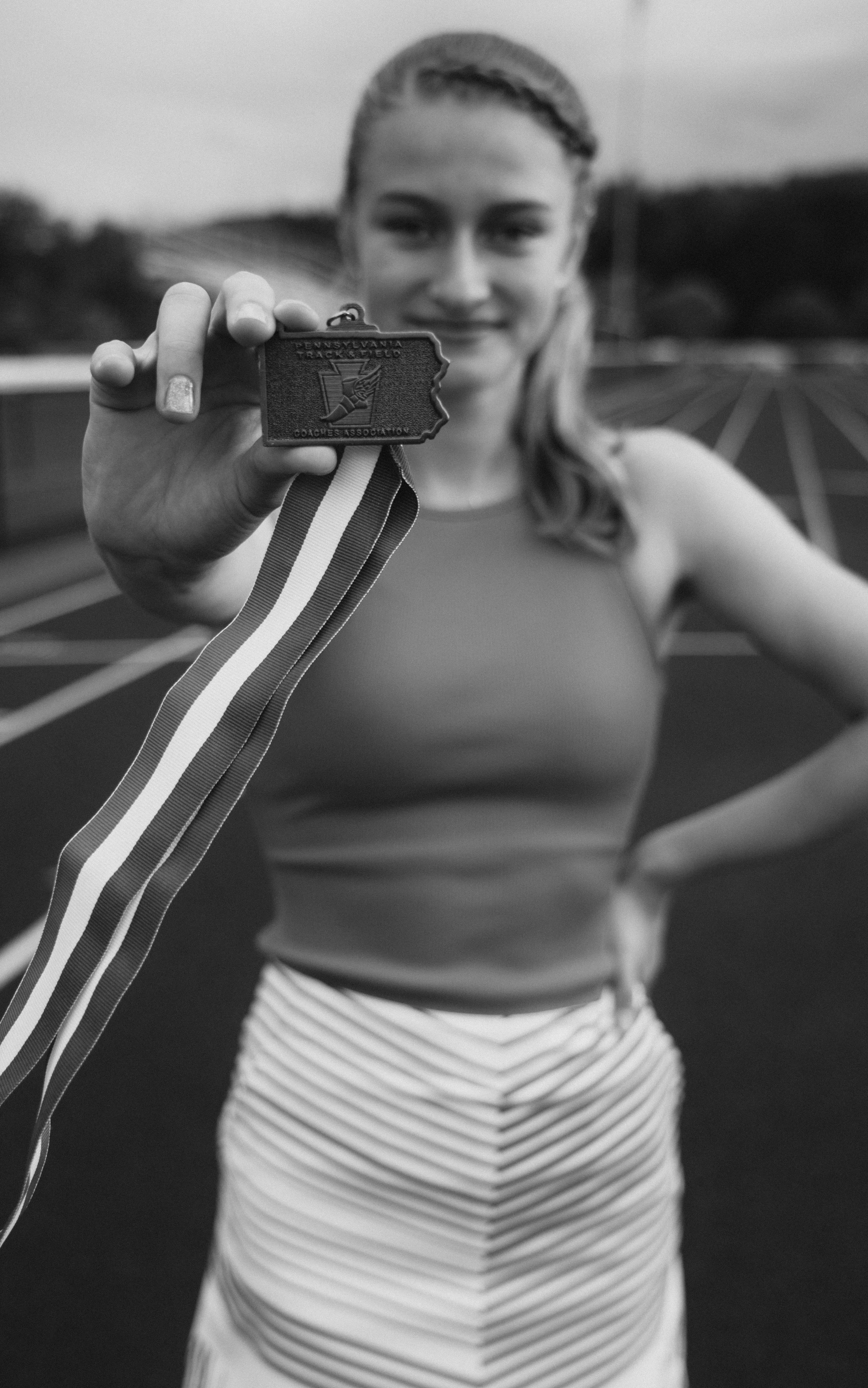 A woman on a running track holding up a medal with the shape of Pennsylvania and the words 'Pennsylvania Track & Field Coaches Association'.