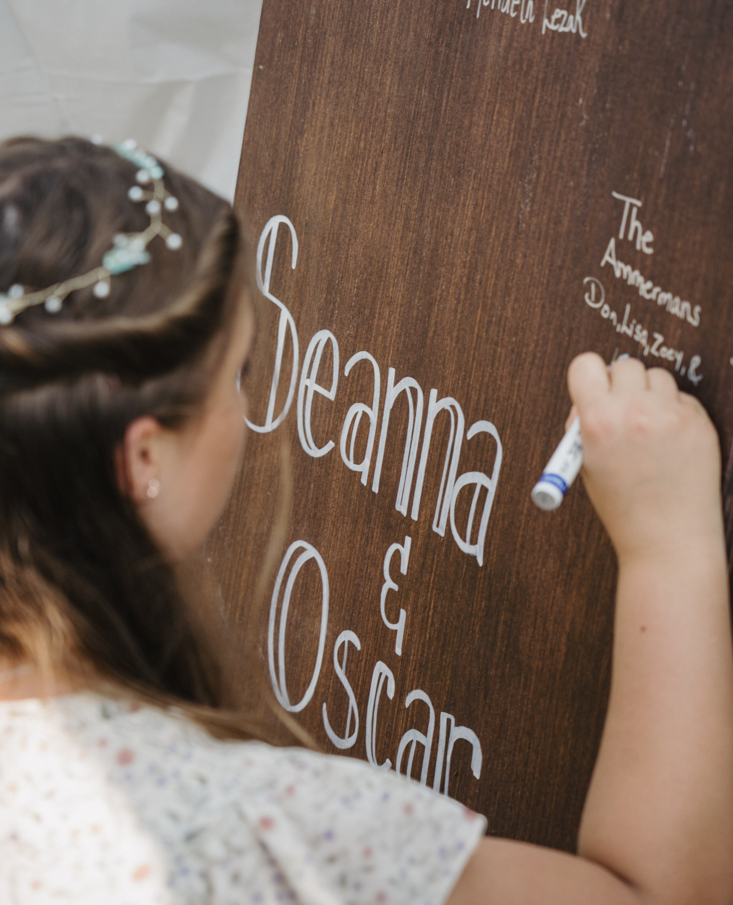 A girl writing on a large wooden sign with white marker, creating handwritten text that reads 'Dreams & Oscar'. She has brown hair and is wearing a floral dress with a floral headband.
