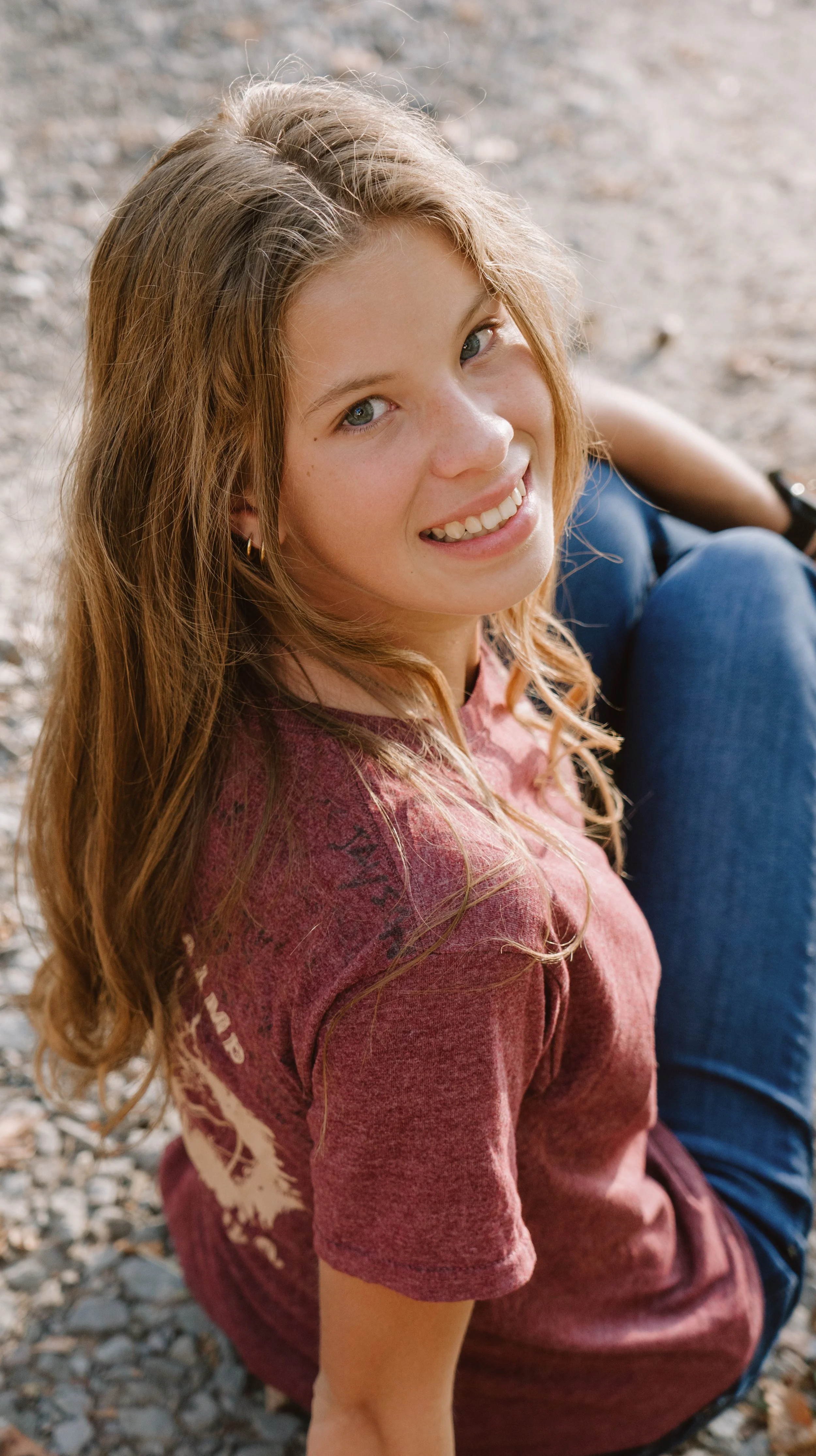 A young woman with long, wavy, reddish hair sitting on a rocky ground outdoors, smiling and looking up at the camera.