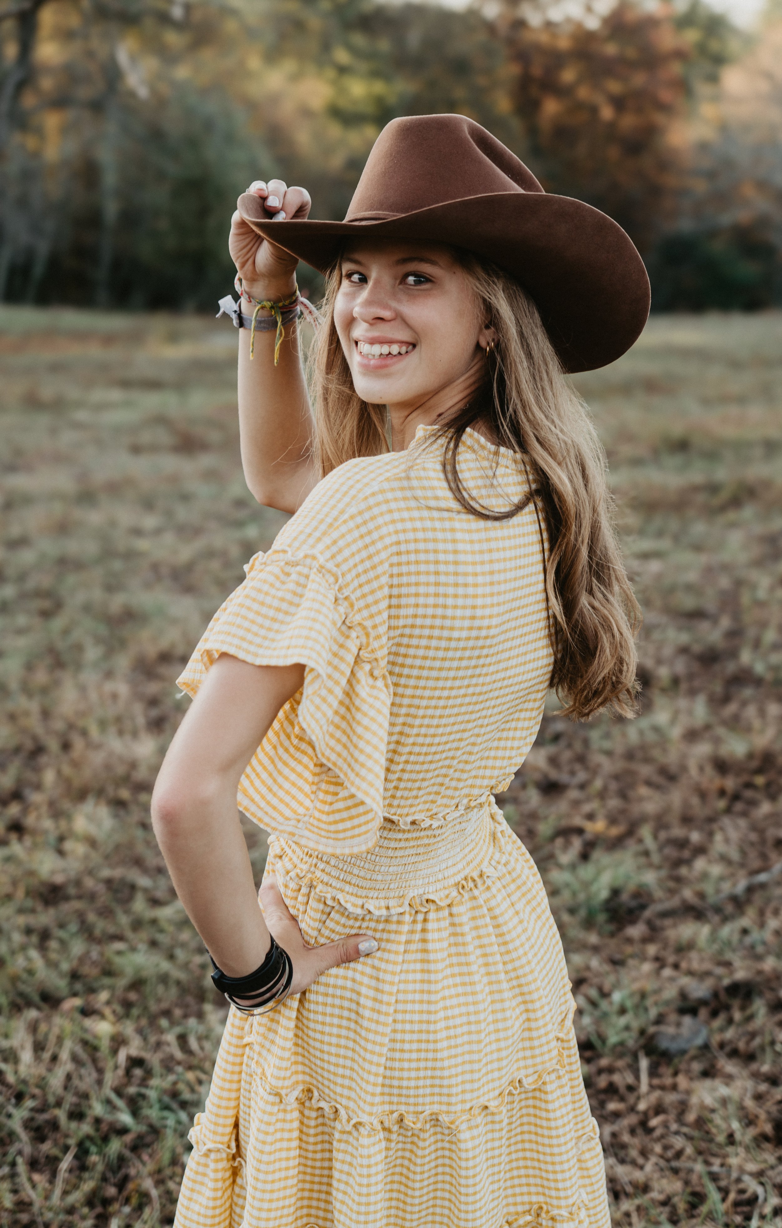 Young woman wearing a yellow checkered dress and brown hat, smiling outdoors in a field with autumn trees in the background.