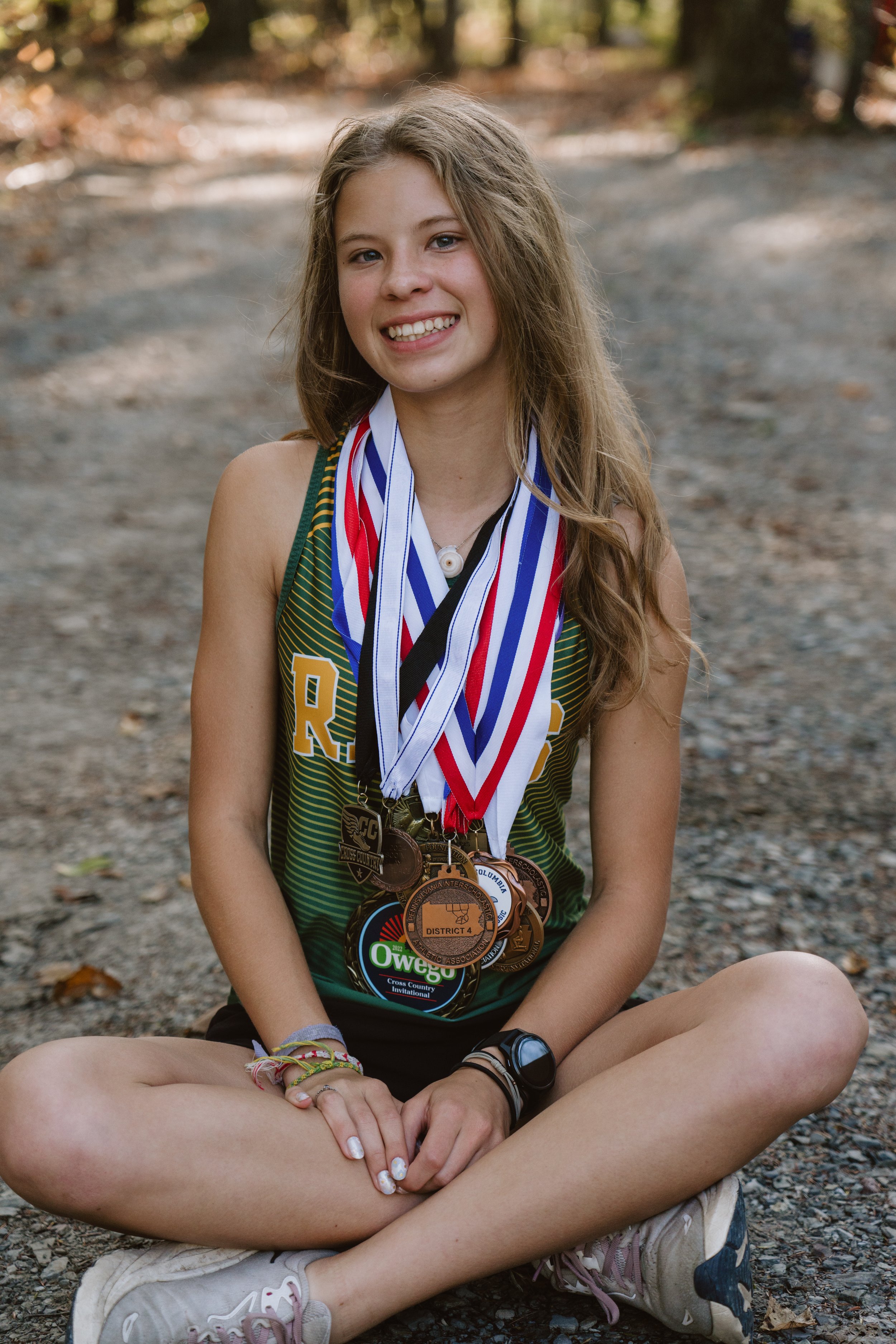 Young girl sitting cross-legged on a gravel path outdoors, smiling, wearing a green athletic jersey with medals hanging around her neck, and adorned with bracelets and a watch.