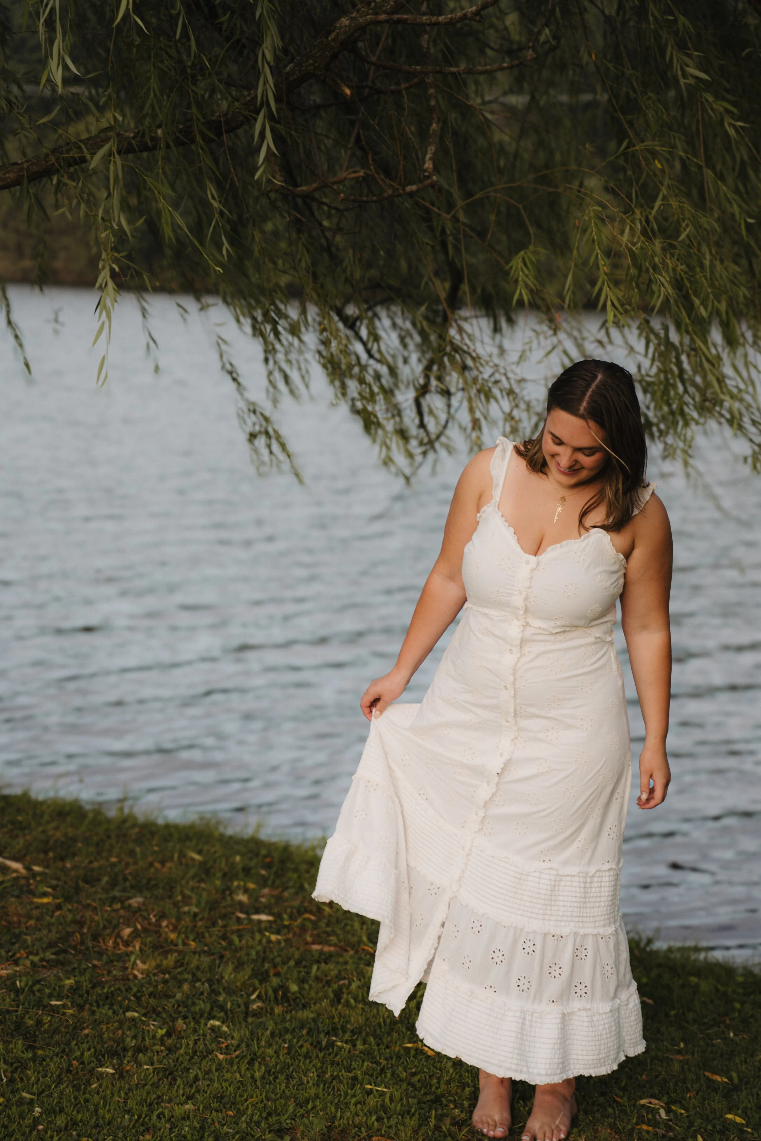 A woman in a white dress standing barefoot near a lake, looking down and smiling, with tree branches and leaves above her.