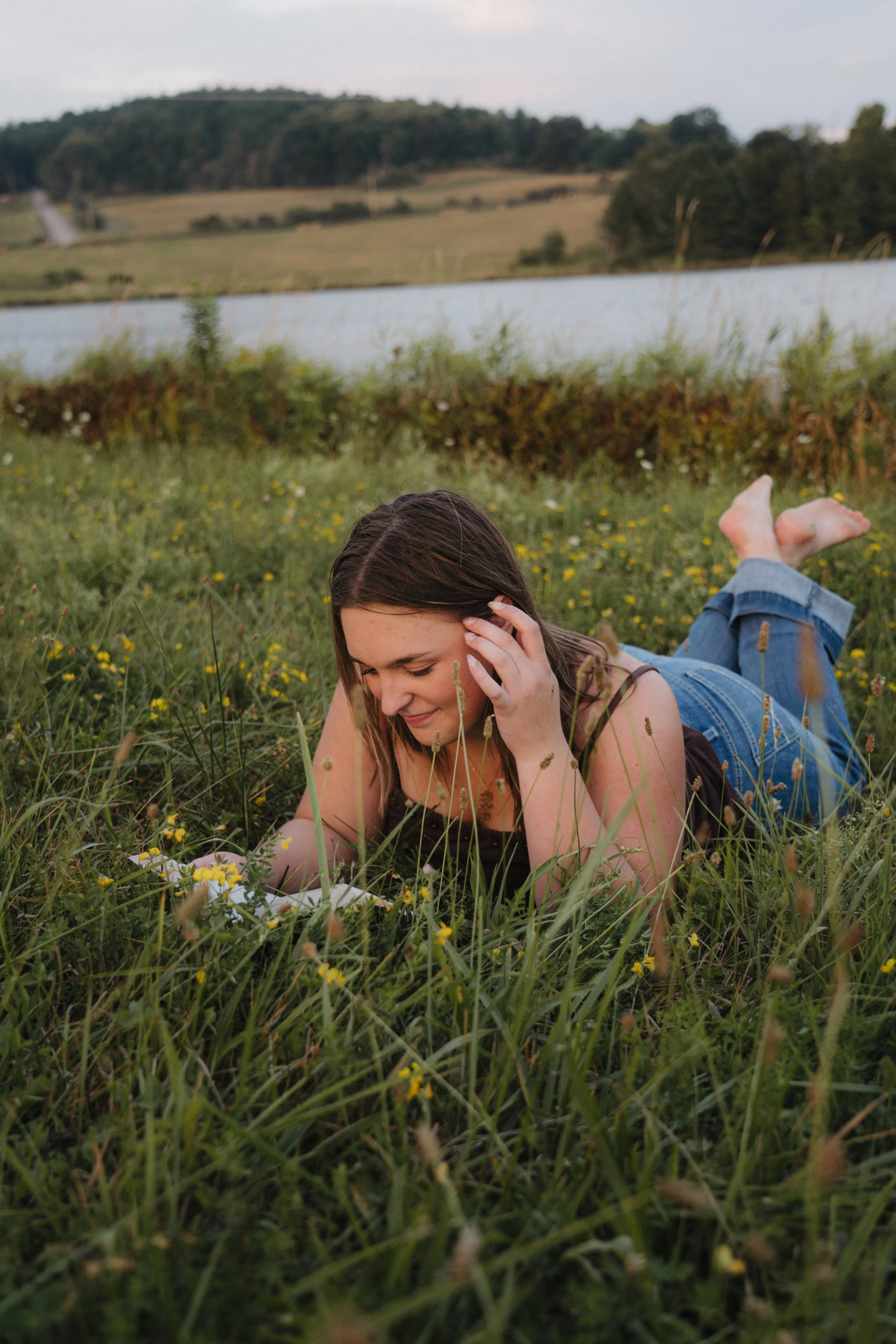 A young woman lying on her stomach in a grassy field near a lake, reading a book and enjoying the outdoors during sunset.