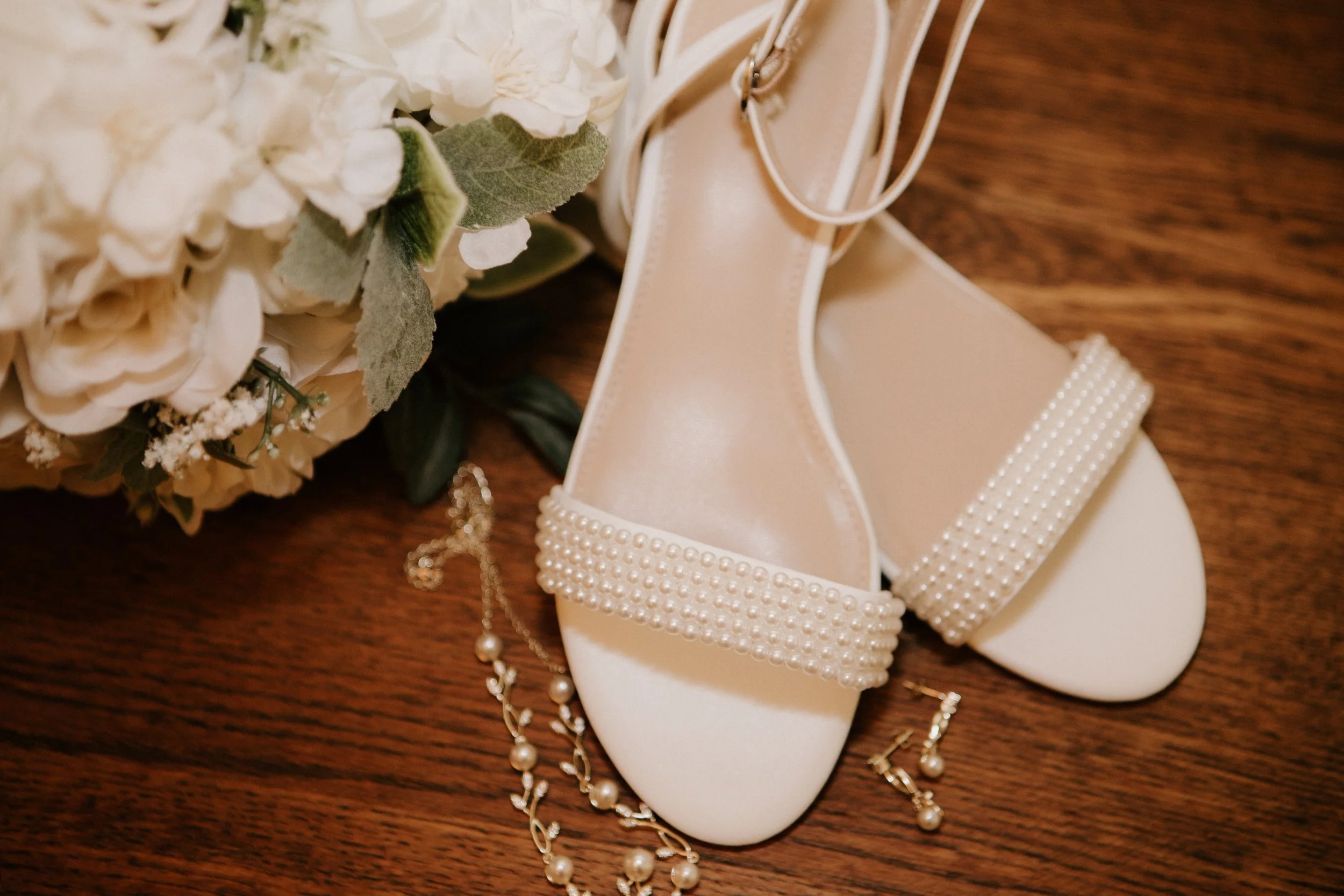 White wedding shoes decorated with pearls, beige flowers, and jewelry on a wooden surface.