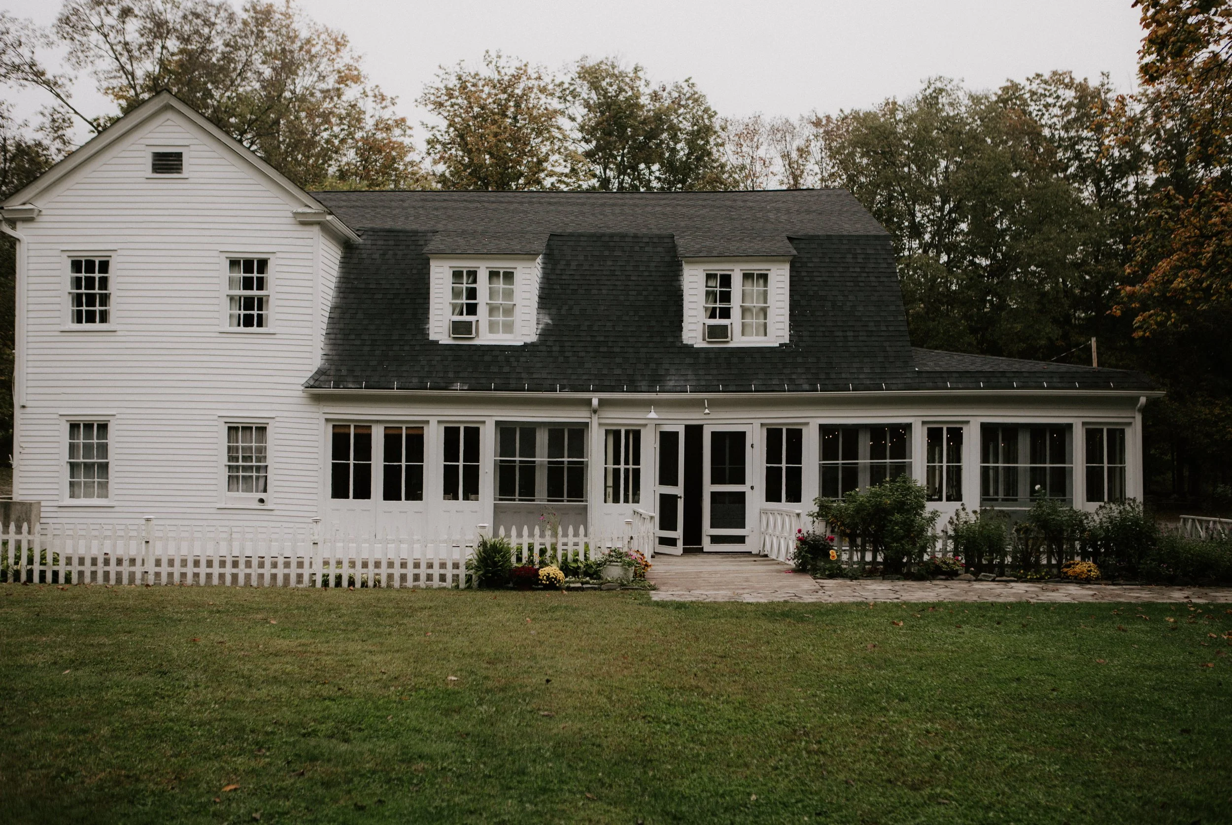 A white two-story house with a porch and a black roof, surrounded by a lawn and some trees in the background.