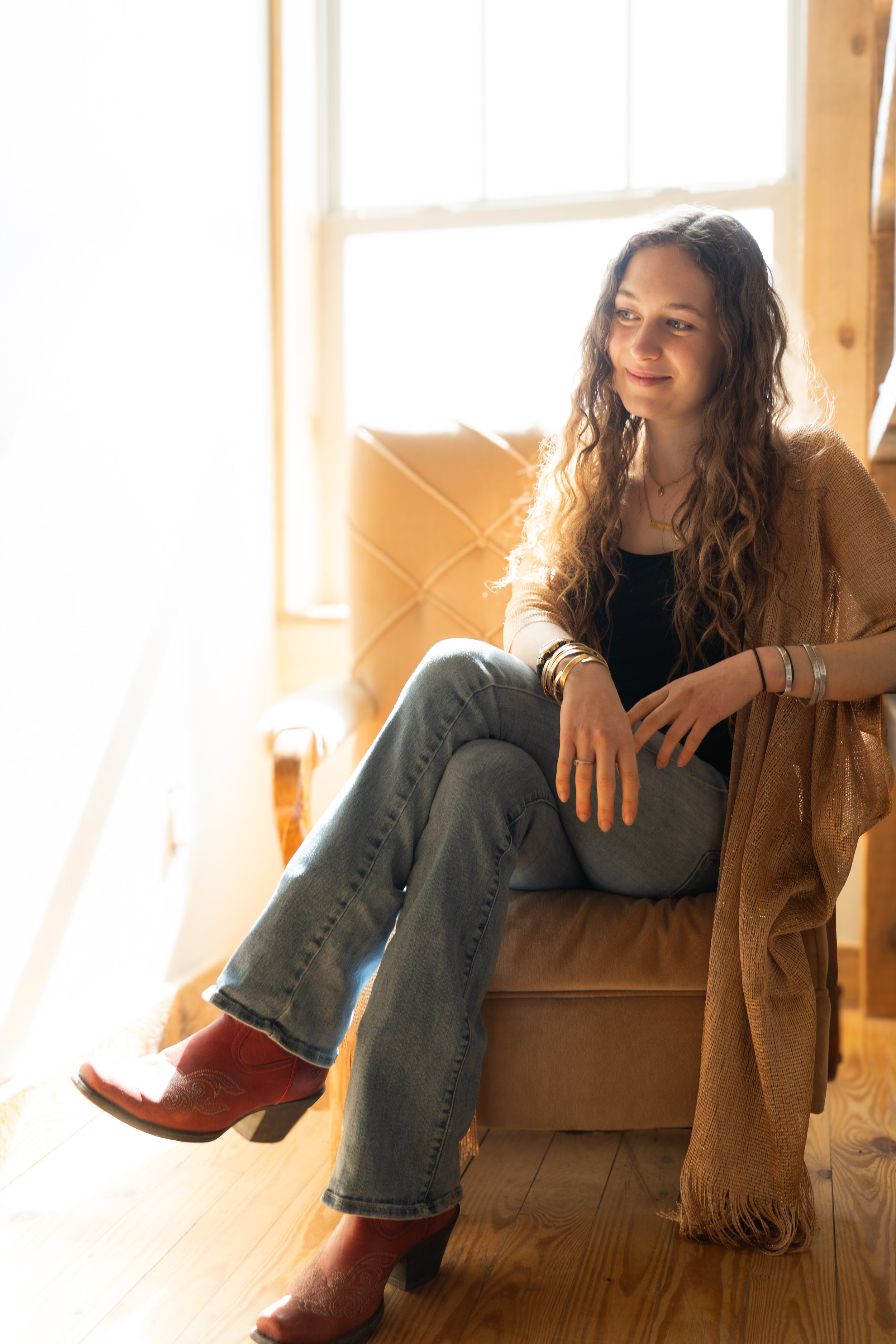 A young woman with long curly hair, sitting on a tan armchair near a window, smiling softly, wearing jeans, a black top, and red cowboy boots, with layered bracelets and a necklace.