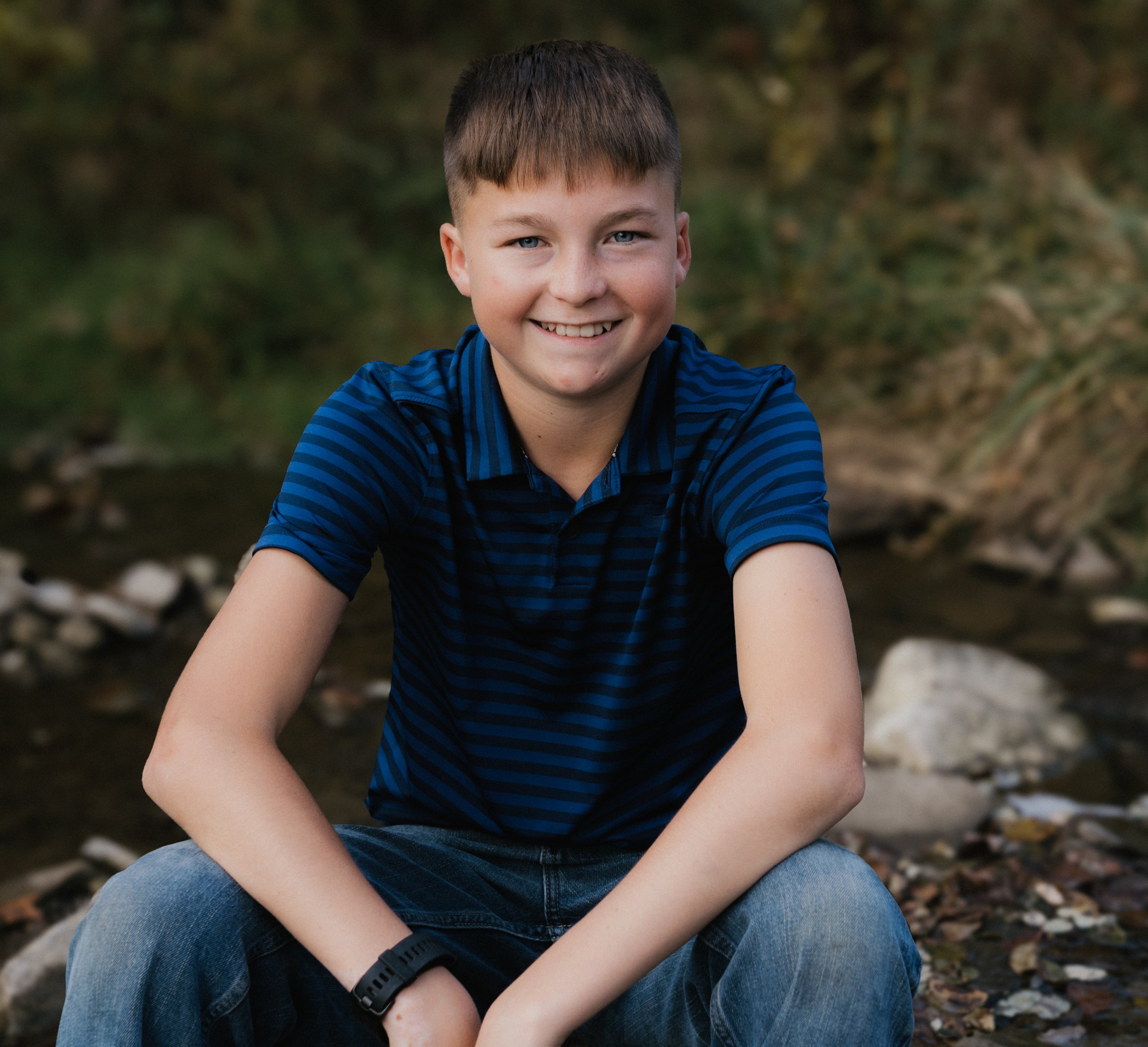 A smiling young boy sitting outdoors near a stream with rocks and greenery in the background.