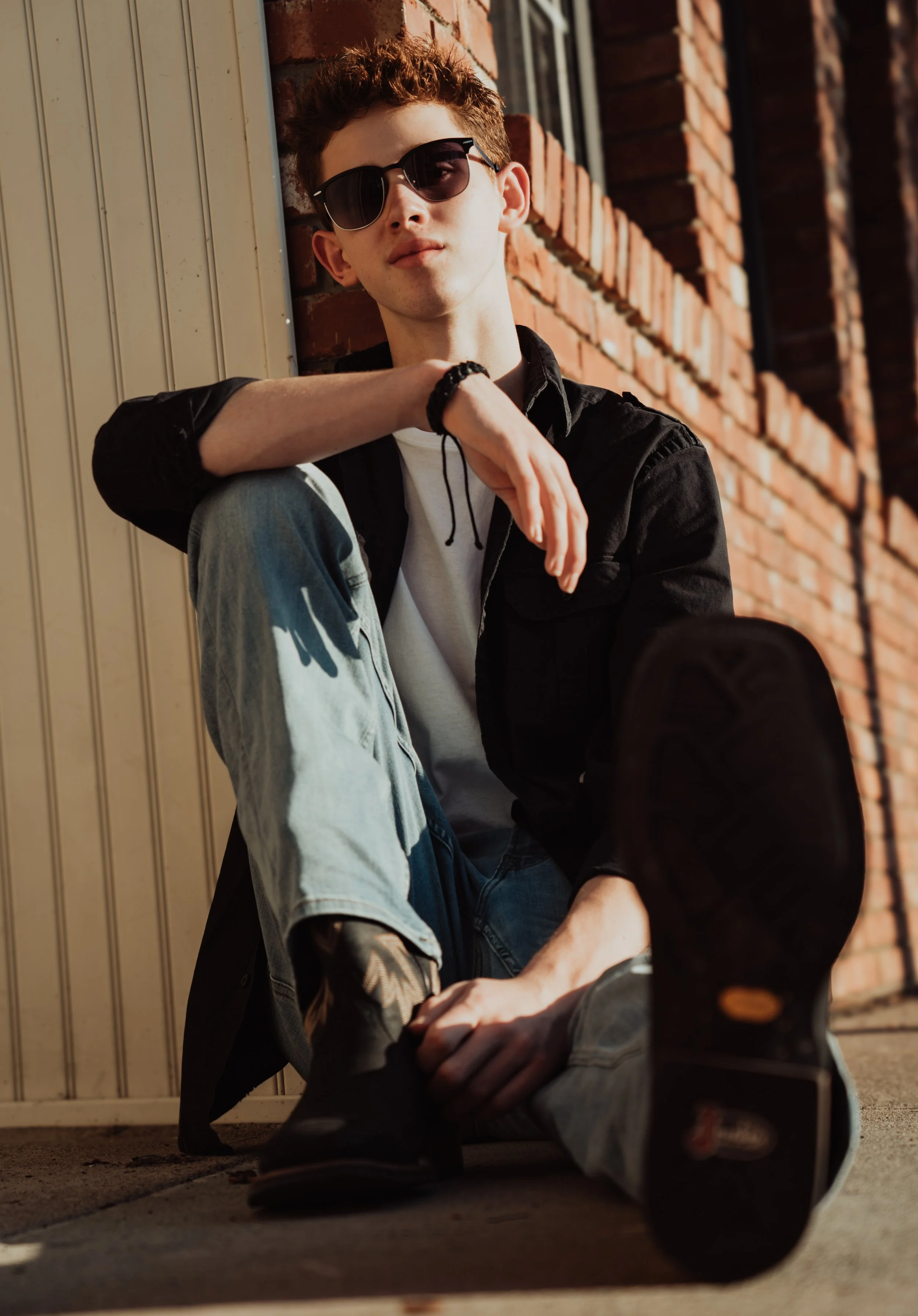 A young man with curly hair wearing sunglasses, a black jacket, a white t-shirt, and ripped light jeans, sitting against a brick wall, looking at the camera with one arm resting on his knee and the other touching his ankle.