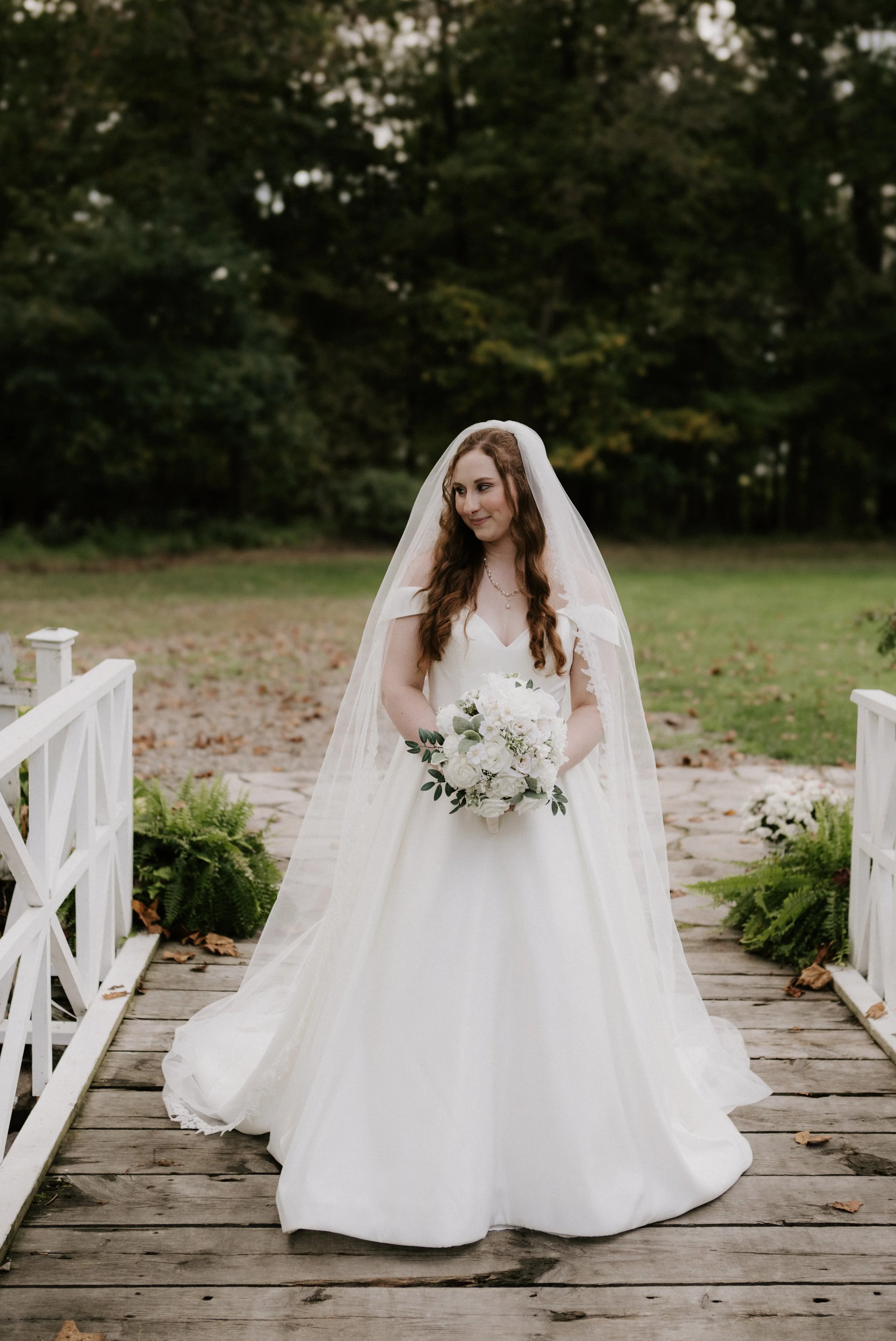A bride in a white wedding gown and veil holding a bouquet of white flowers standing on a wooden bridge with white railings, surrounded by green plants and trees in a park setting.