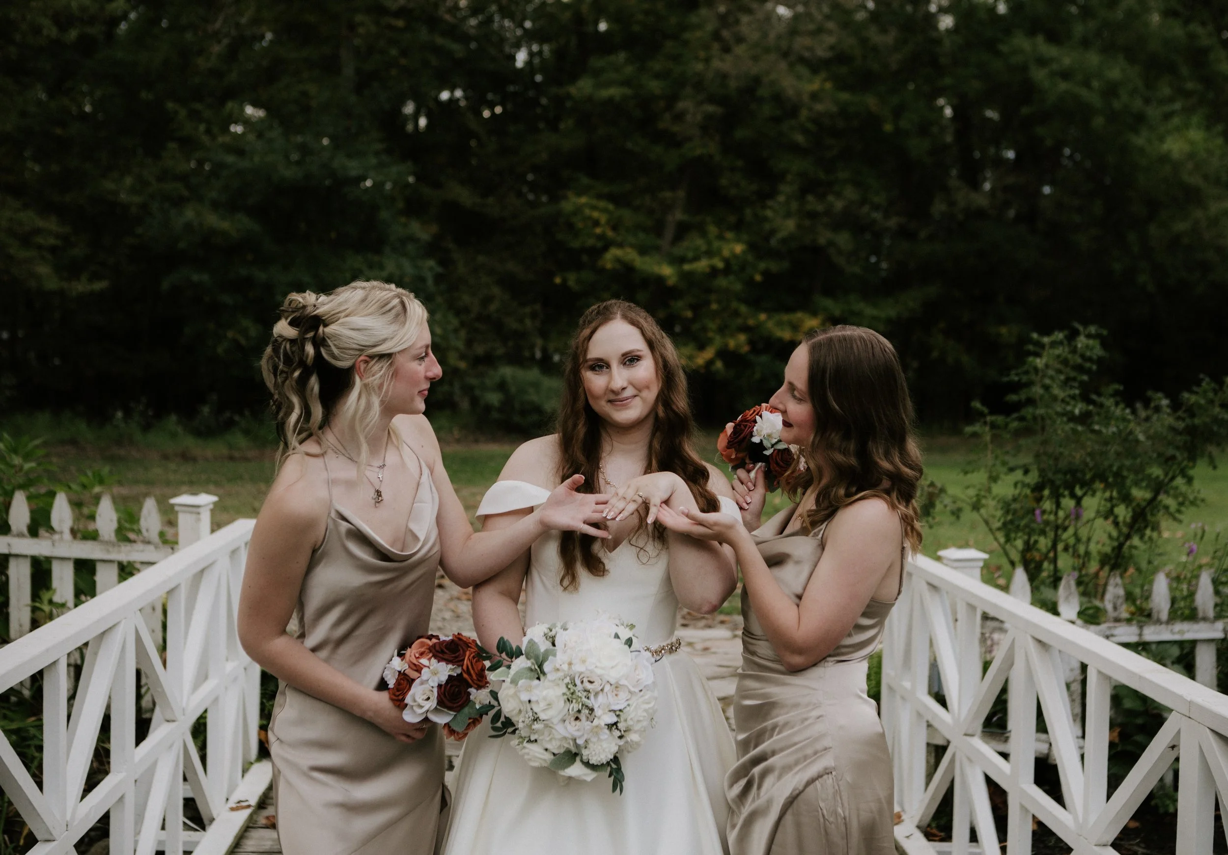 Three women at a wedding, one bride in the center in a white dress holding a bouquet, flanked by two bridesmaids in matching champagne dresses holding bouquets, on a white wooden bridge with greenery in the background.