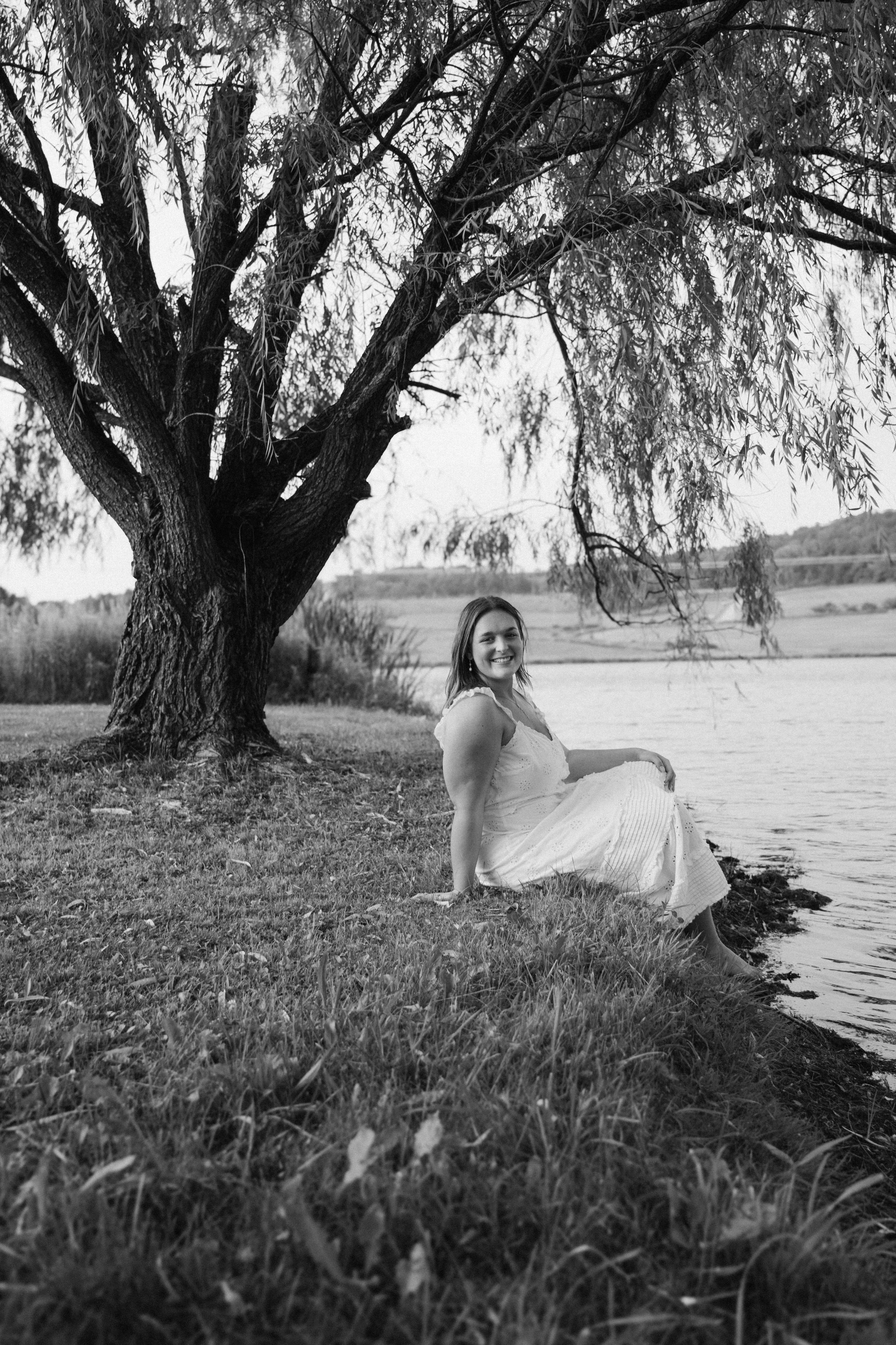 A woman in a white dress sitting by a riverbank beneath a large tree, smiling at the camera, in black and white.