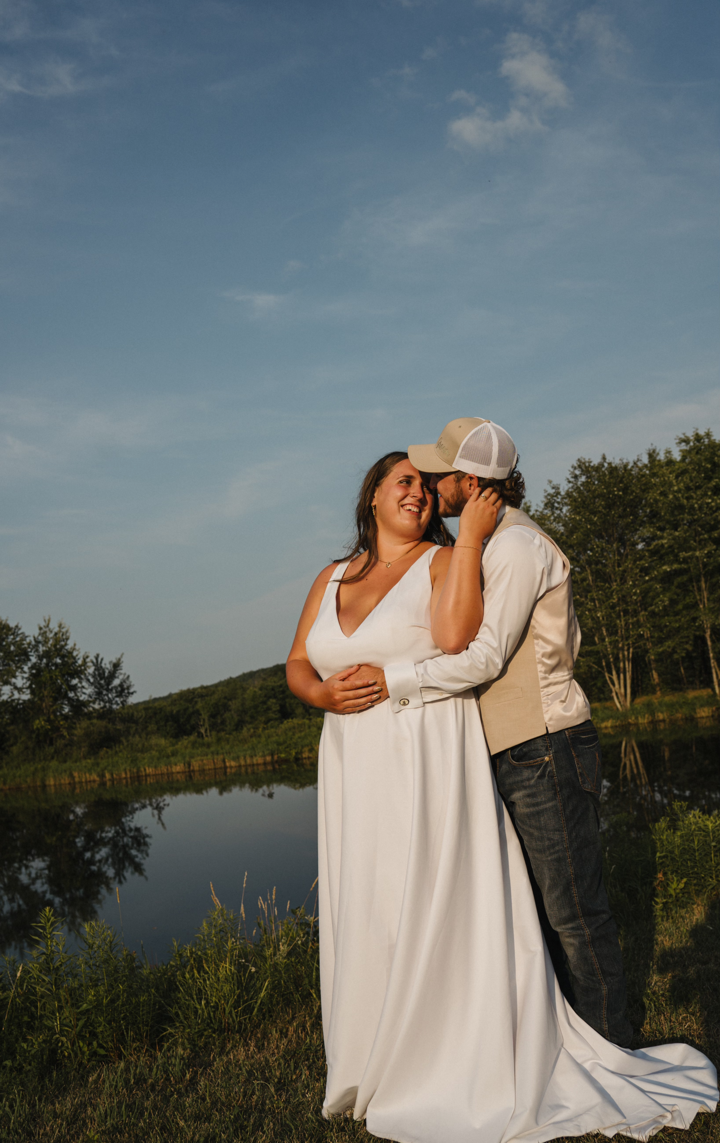 A couple dressed in wedding attire embracing outdoors near a lake, with trees and hills in the background, under a partly cloudy sky.