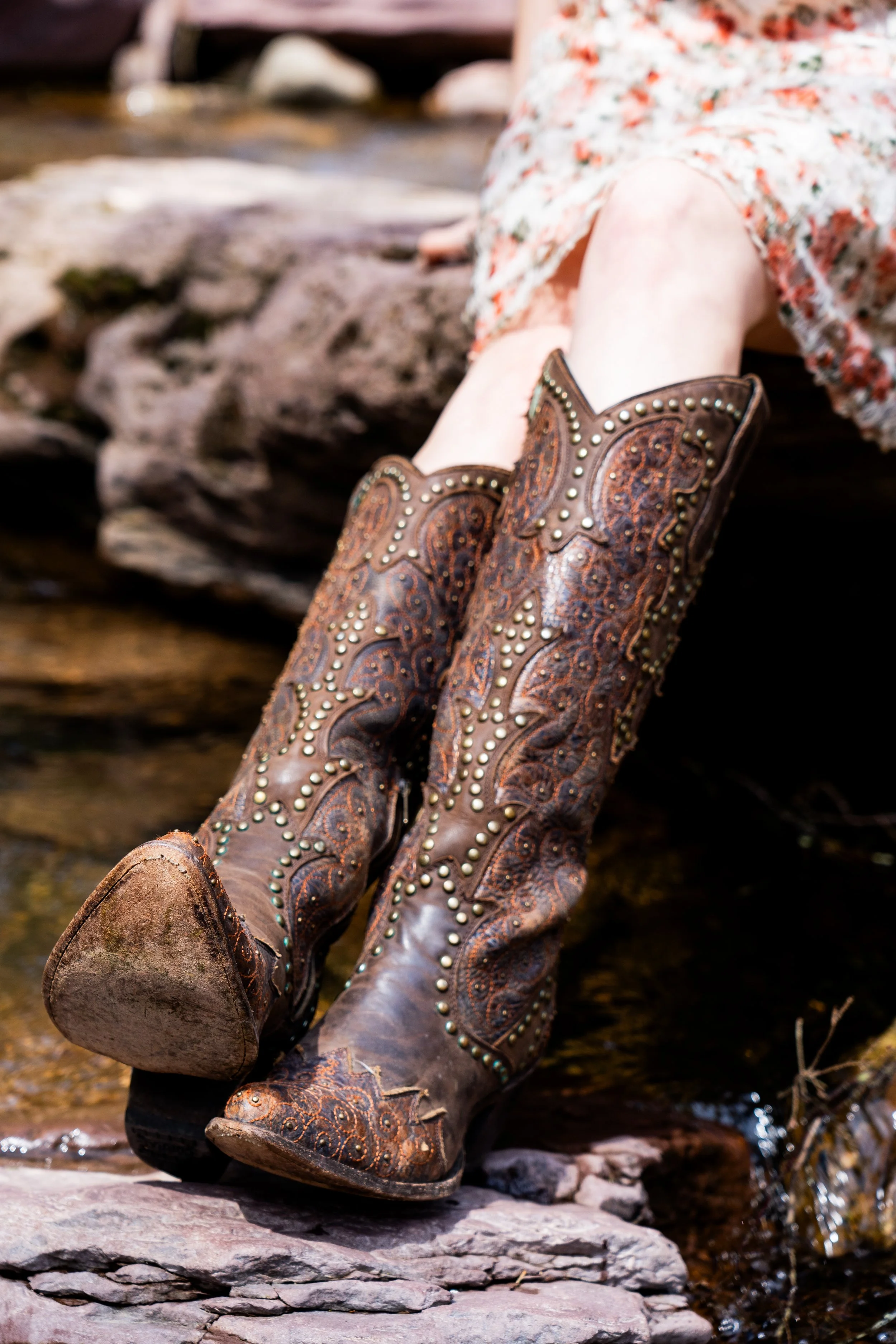 A close-up of a person's legs wearing brown decorated cowboy boots while sitting on rocks near a stream.
