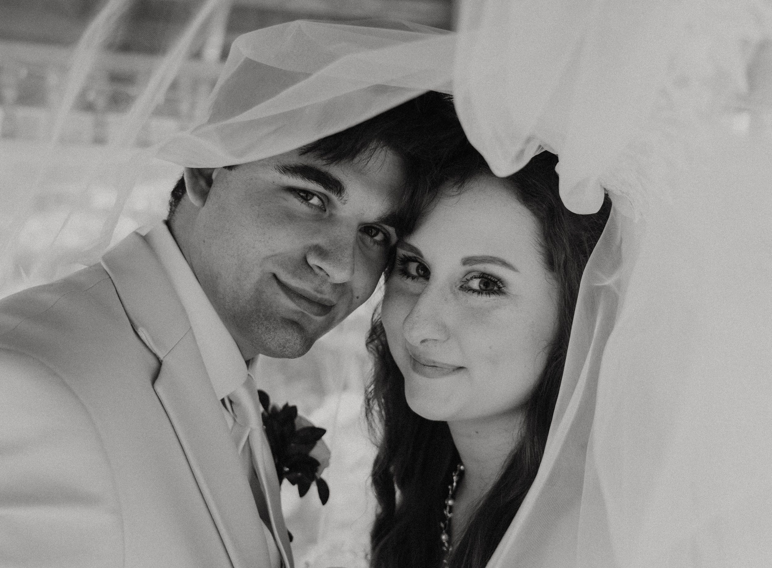 Black and white photo of a man and a woman close together, under a veil, smiling at the camera.