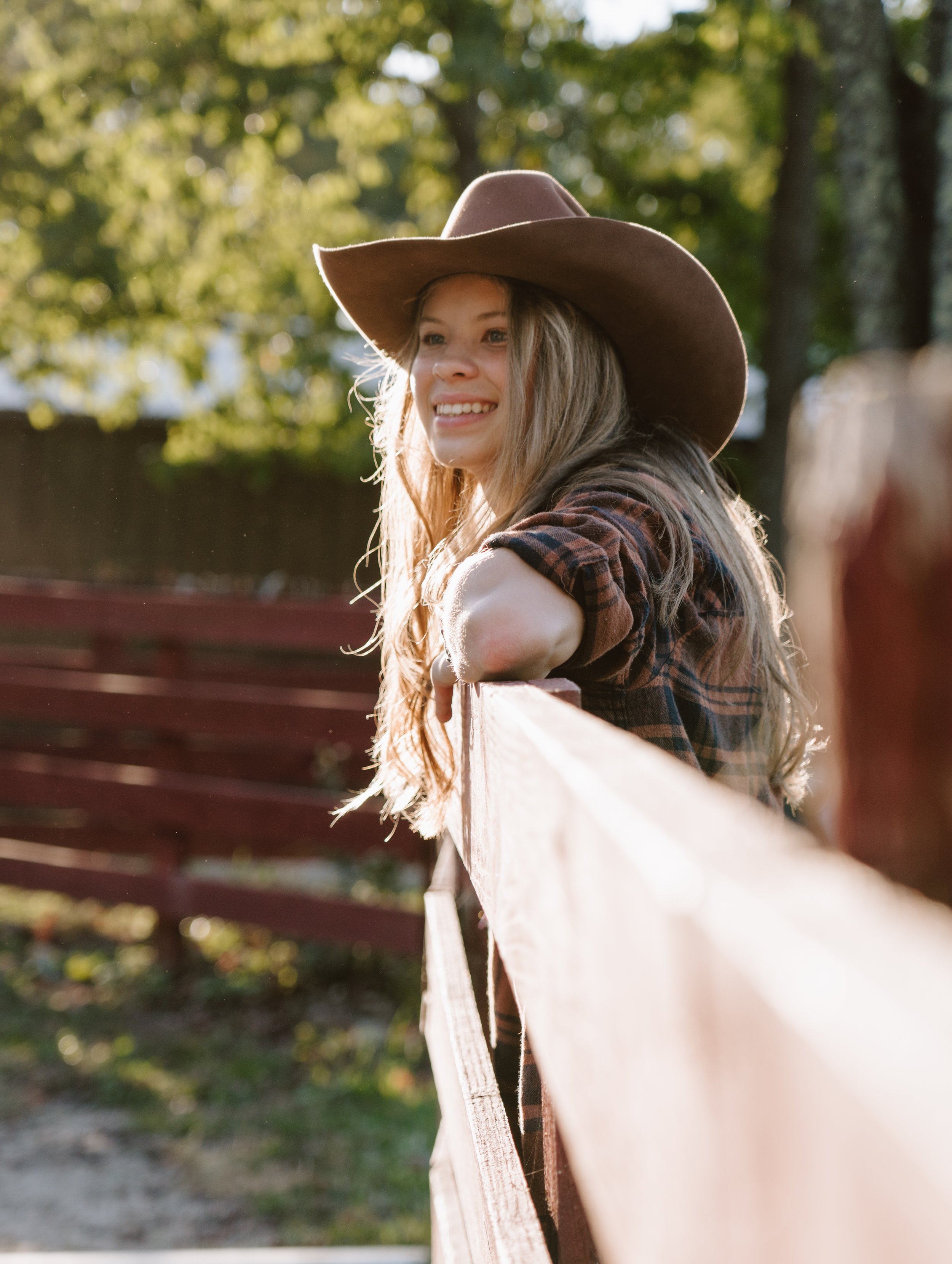 A young woman with long blonde hair wearing a cowboy hat and plaid shirt, leaning on a wooden fence outdoors in sunlight.