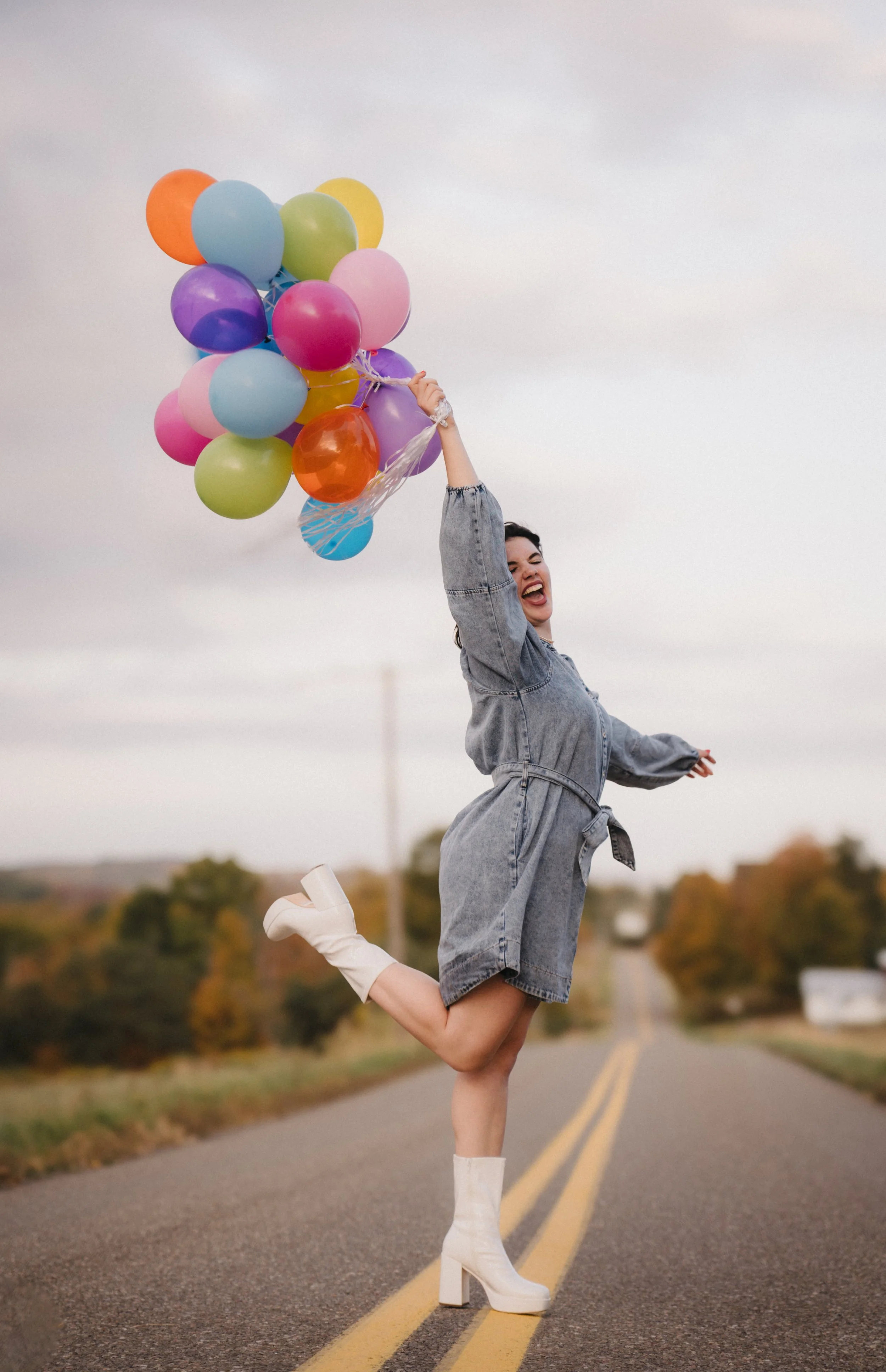 A woman in a denim dress and white high-heeled boots joyfully holding a bunch of colorful balloons while jumping on an empty rural road with trees in the background.