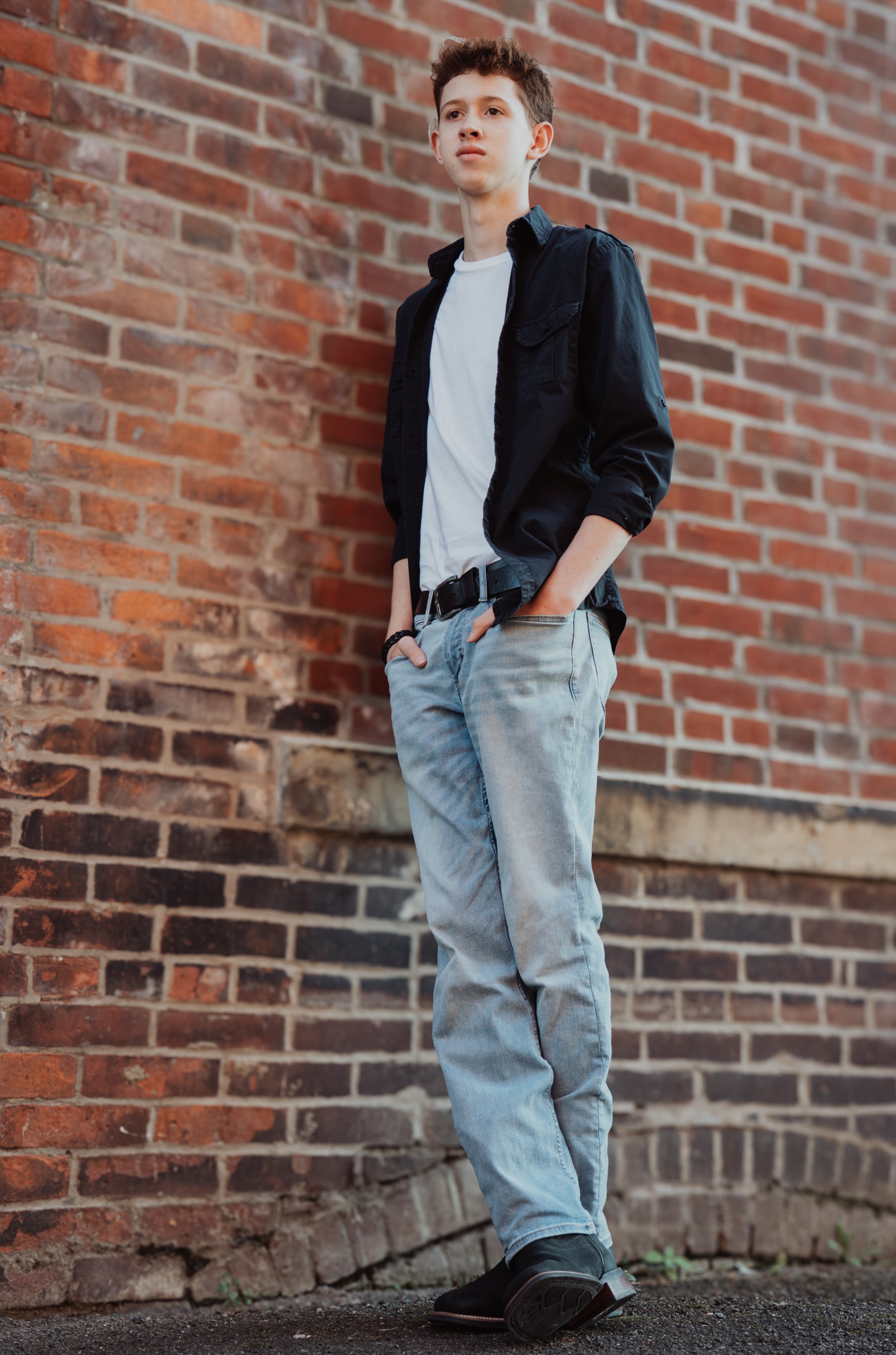 A teenage boy standing against a red brick wall, wearing a black jacket over a white T-shirt and light blue jeans with hands in pockets.