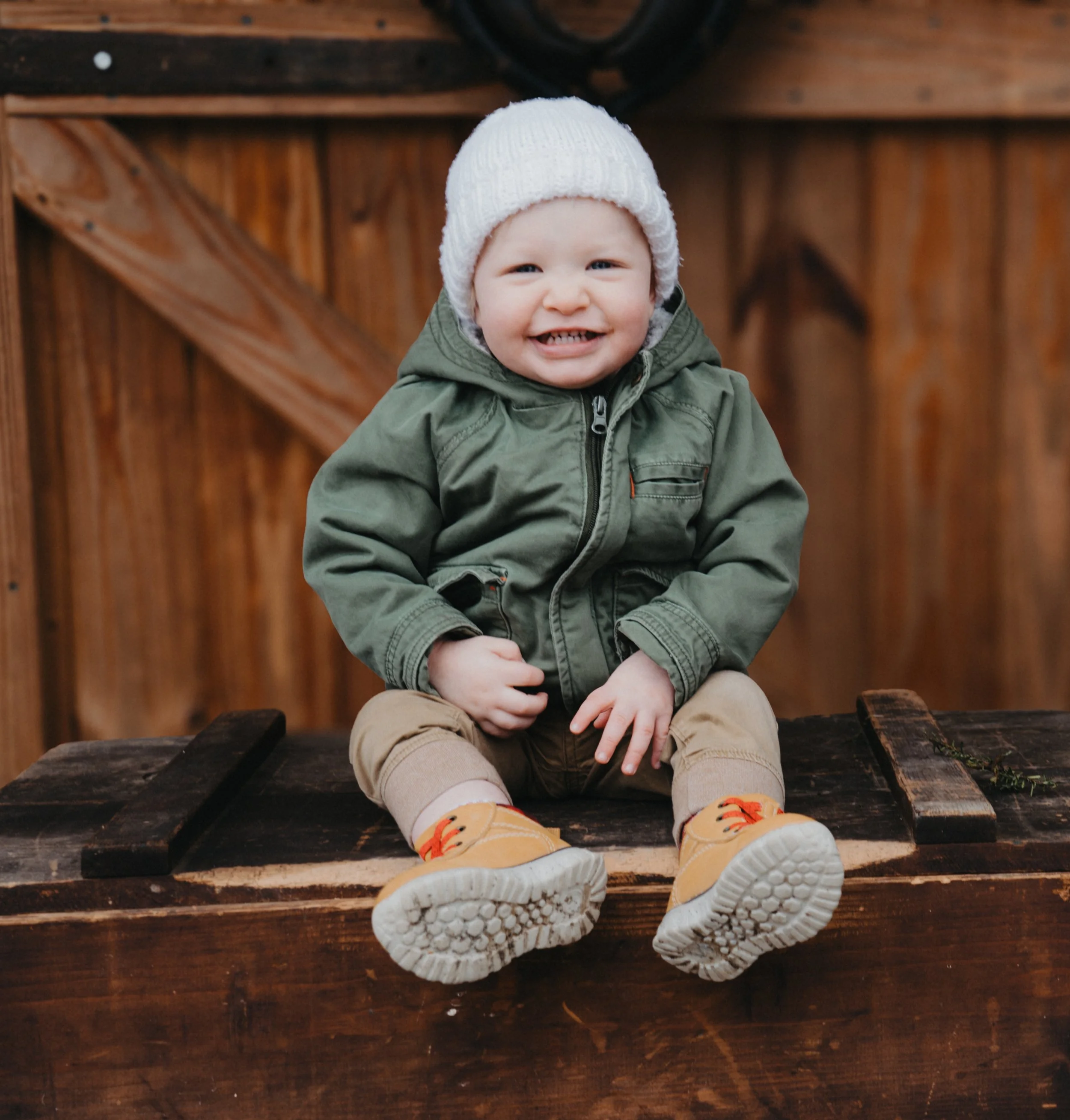 A smiling young child with a white knit hat, green jacket, tan pants, and yellow shoes sitting on a wooden surface in front of a wooden fence.