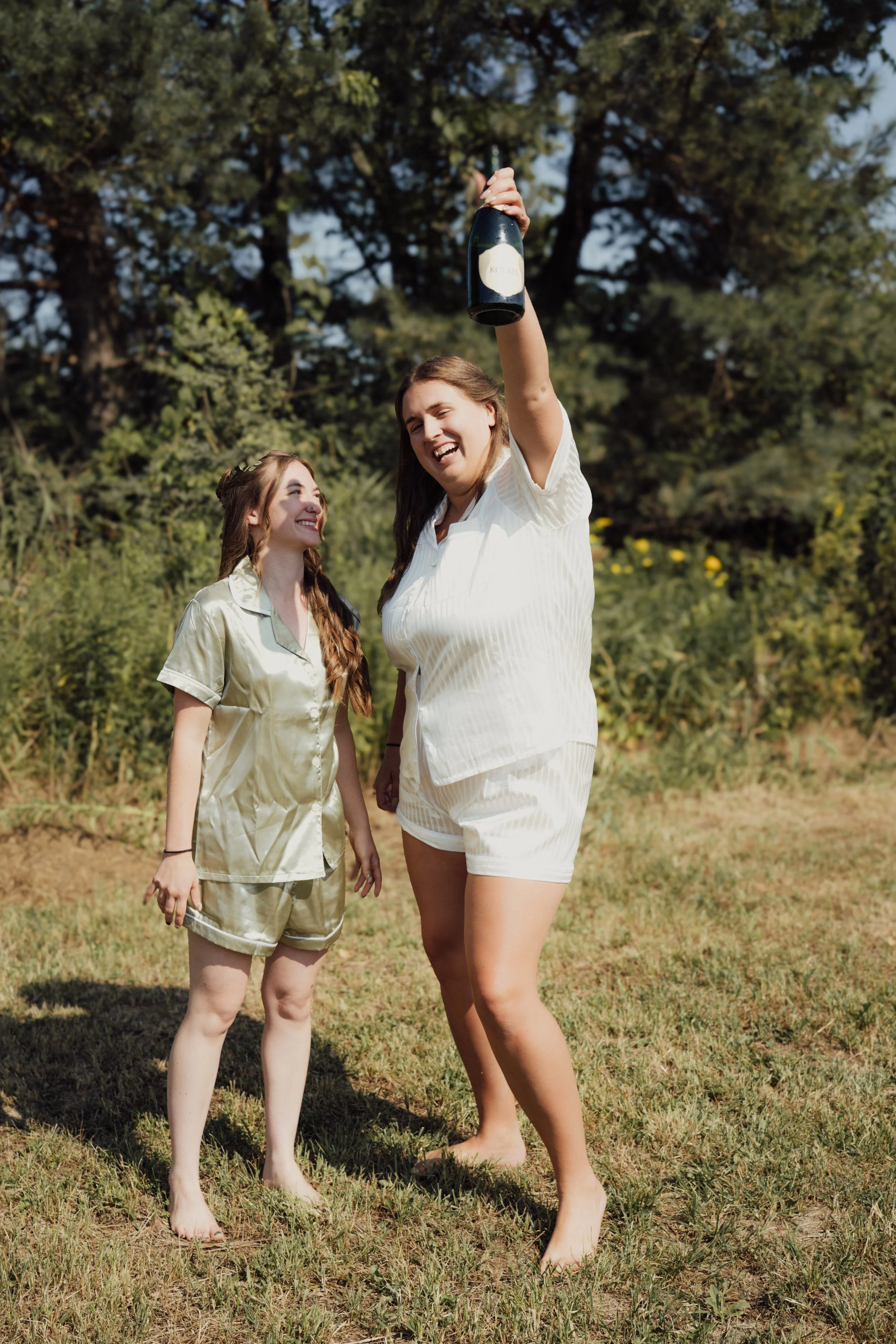 Two women in pajamas standing outdoors in a grassy area, celebrating with a bottle of champagne, smiling and enjoying a sunny day.