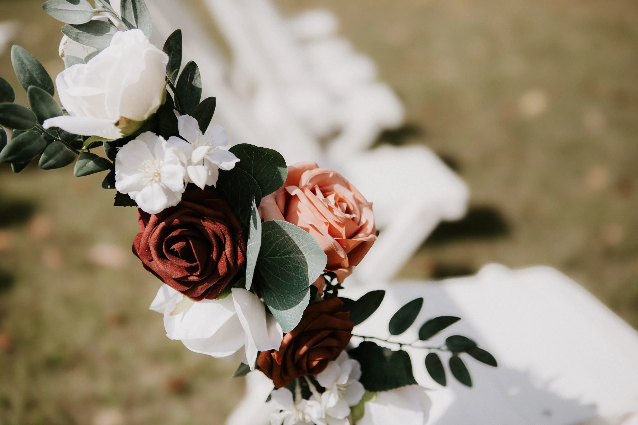Close-up of a floral arrangement with white, pink, and red roses, white flowers, and green foliage on a white bench, outdoors.